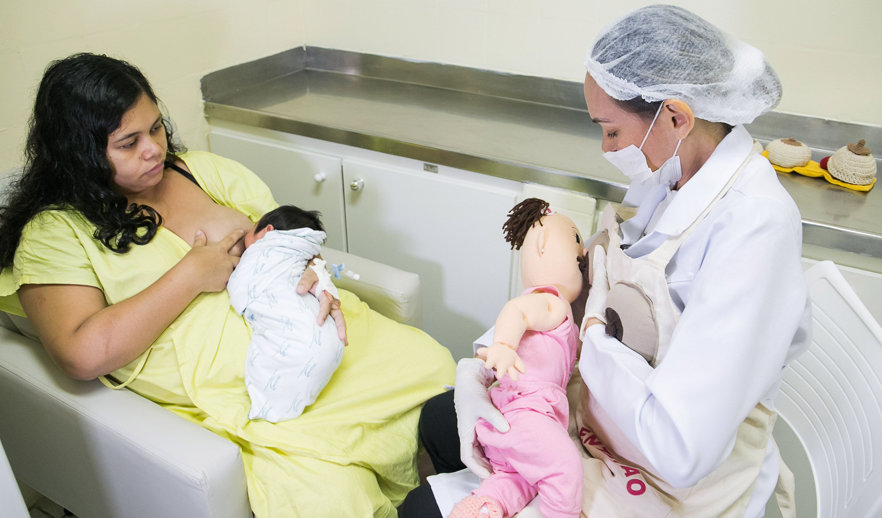 Mulher amamentando e olhando para enfermeira simulando amamentação com boneca