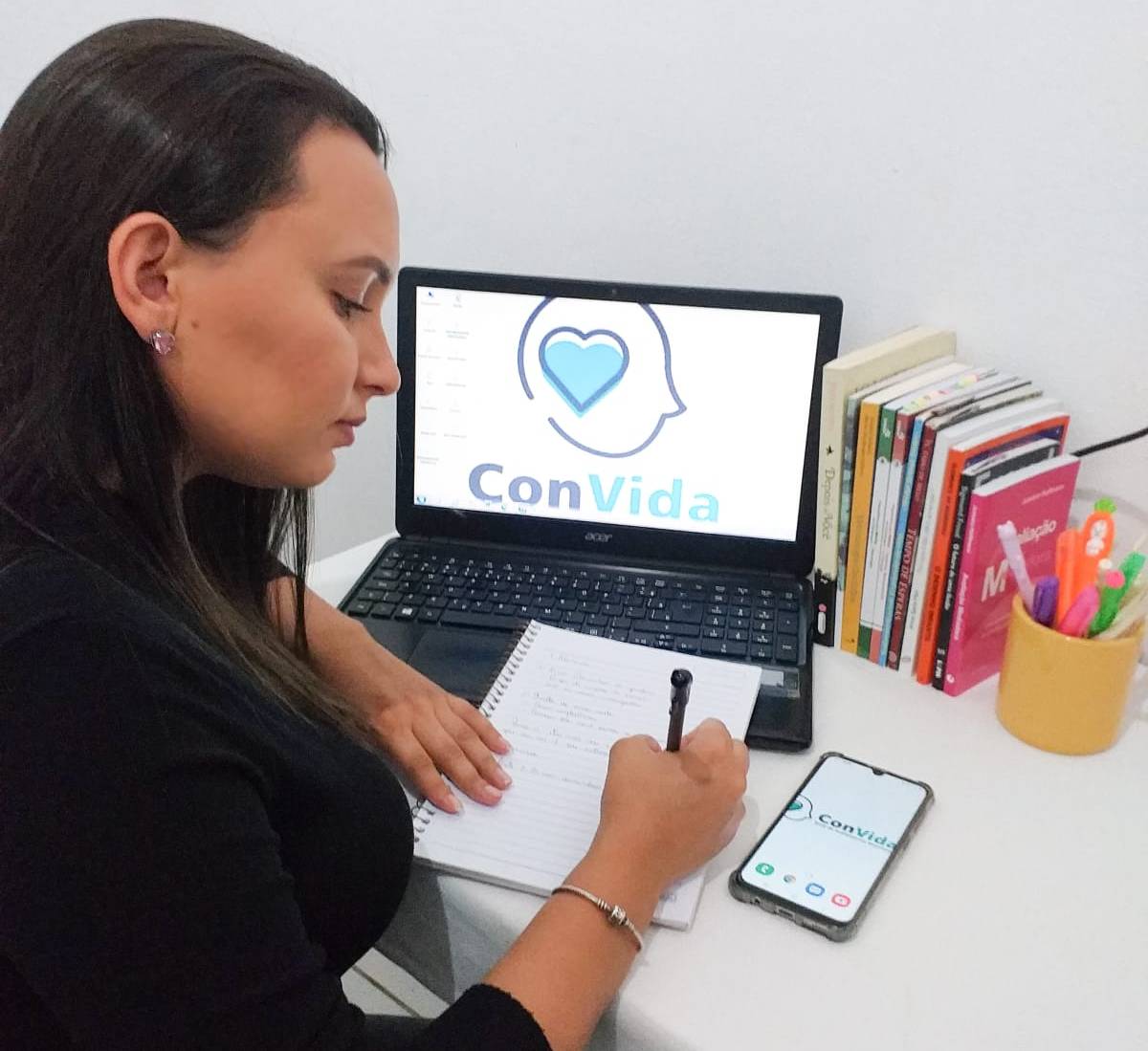psicóloga sentada em frente a computador com a logomarca da ConVida e escrevendo em um caderno