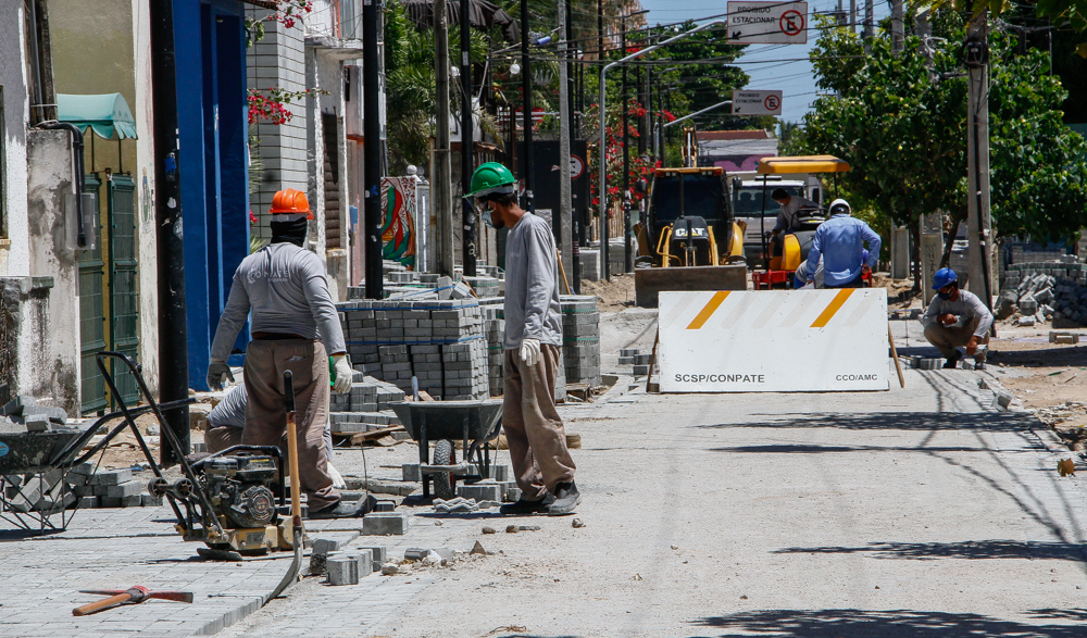 operários trabalhando em rua em obras, recebendo instalação de piso intertravado e placa de obra ao fundo