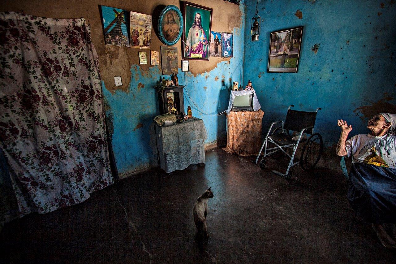 Fotografia no interior de uma casa simples de uma religiosa em Juazeiro do Norte. Nas paredes várias imagens de Jesus e de santos. Uma senhora está sentada na cadeira de balanço no canto direito inferior da foto.