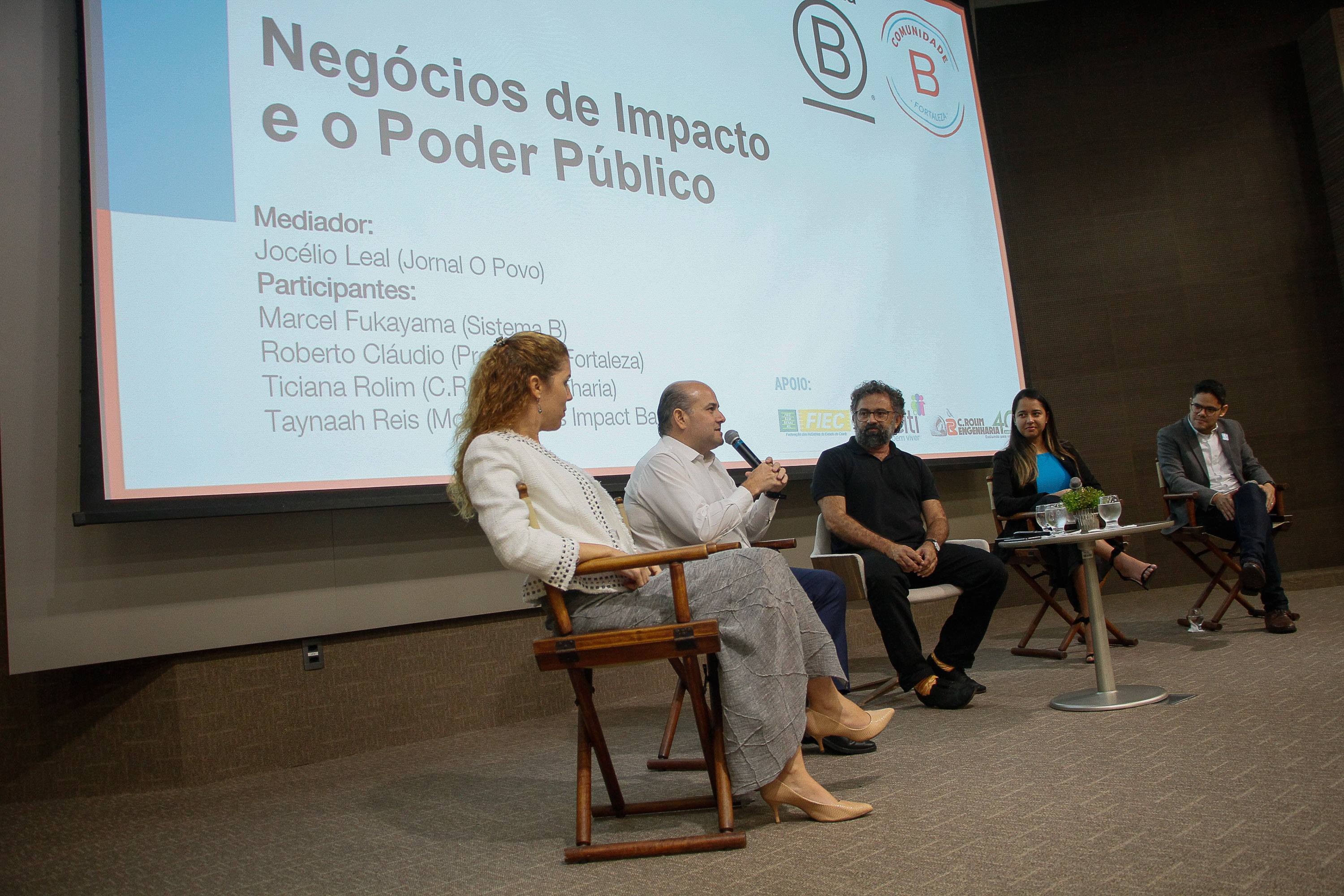 Prefeite em palco de auditório fala ao microfone ao lado de outros palestrantes