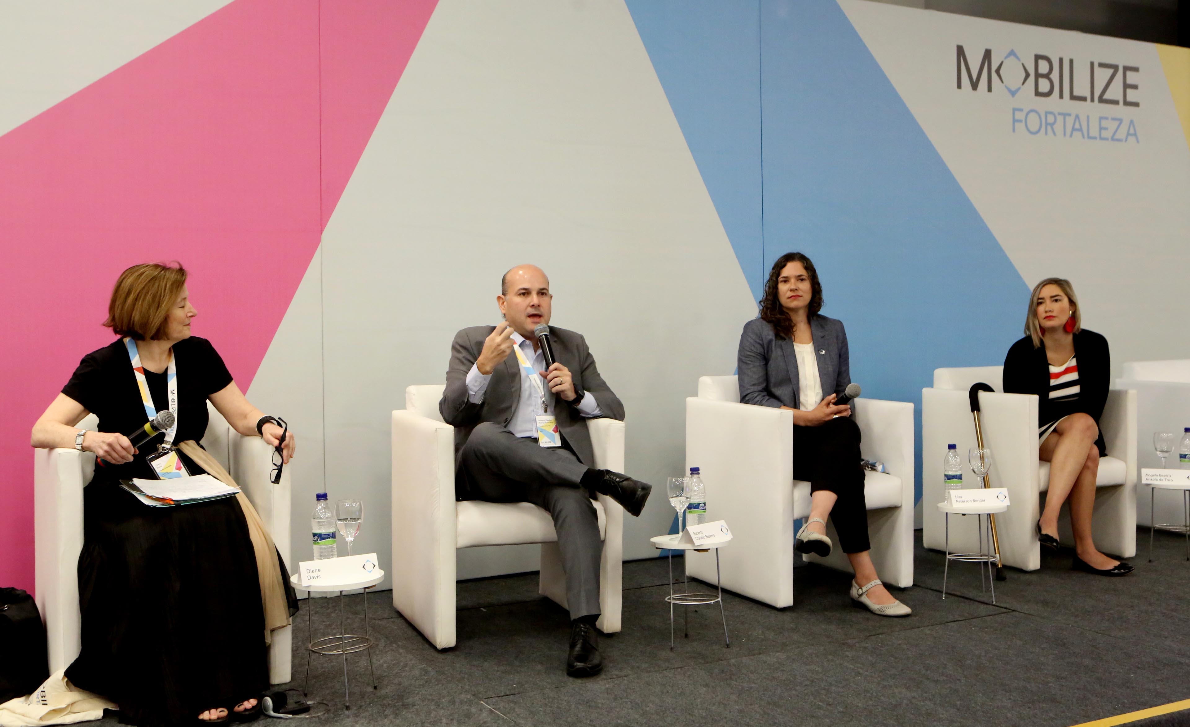 Prefeito sentado em cadeira em cima de palco fala ao microfone