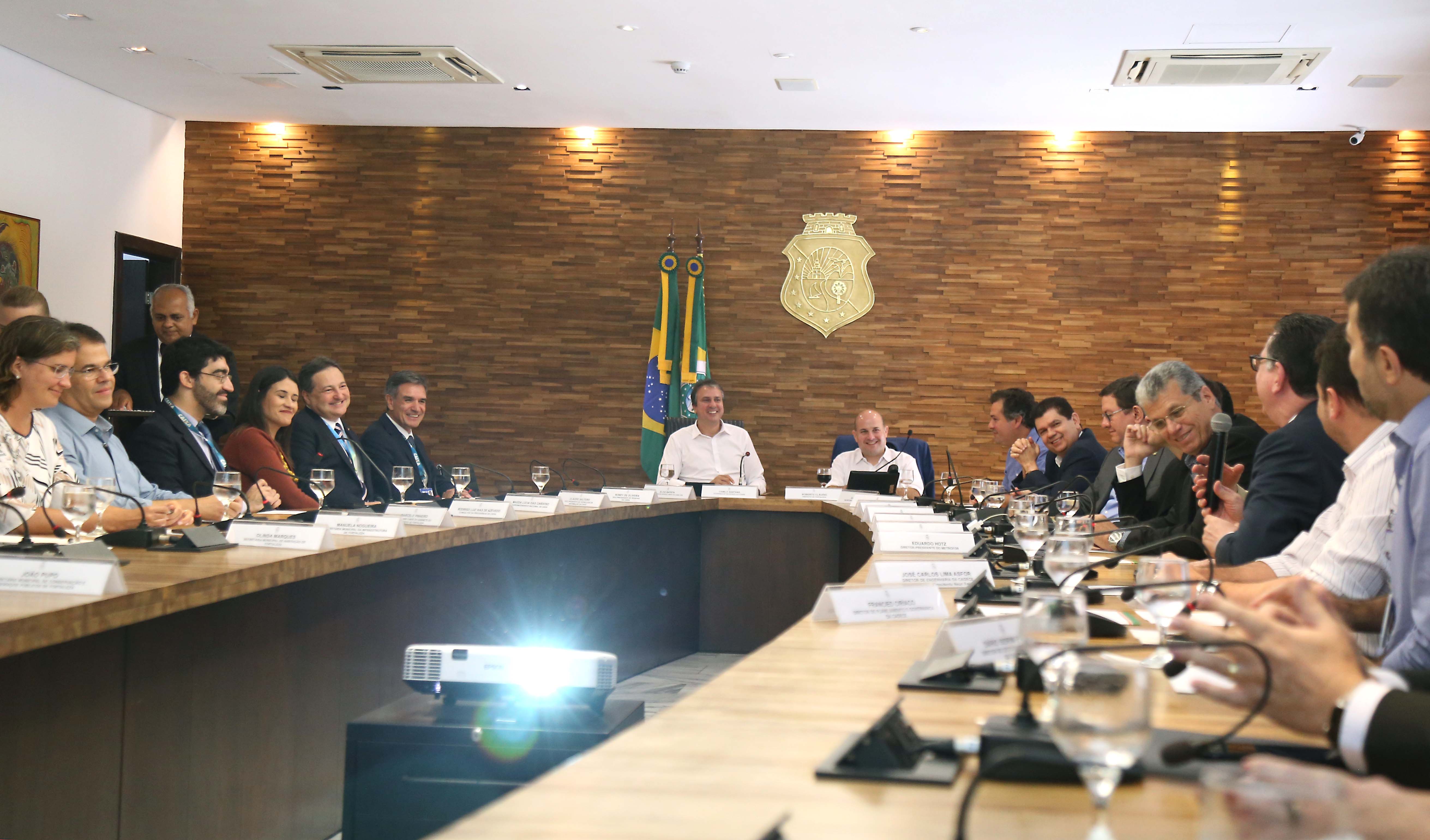 Governador e prefeito sentador ao funo em mesa com várias pessoas