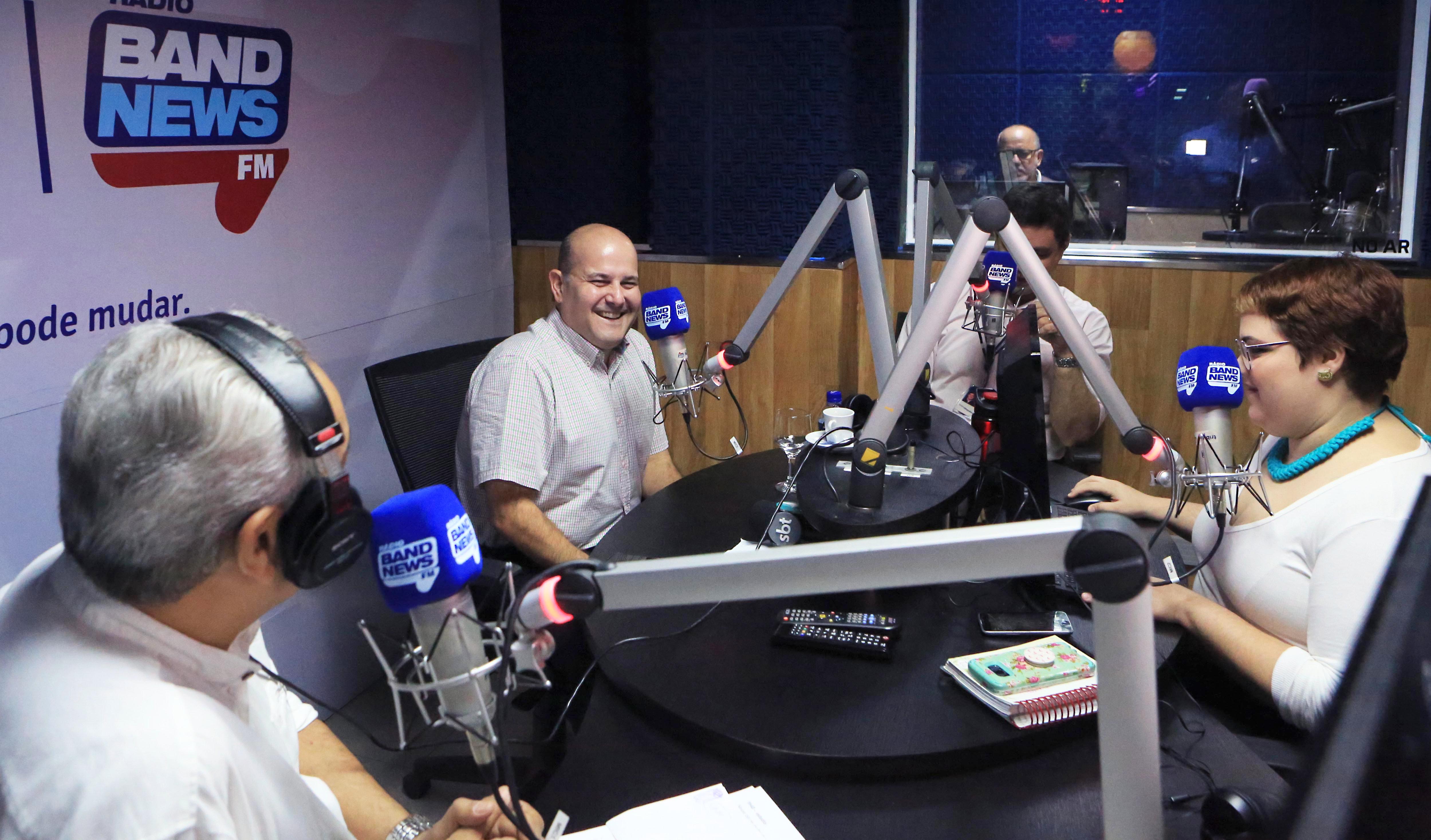 Prefeito sentado à mesa em stúdio de rádio ao lado de jornalistas