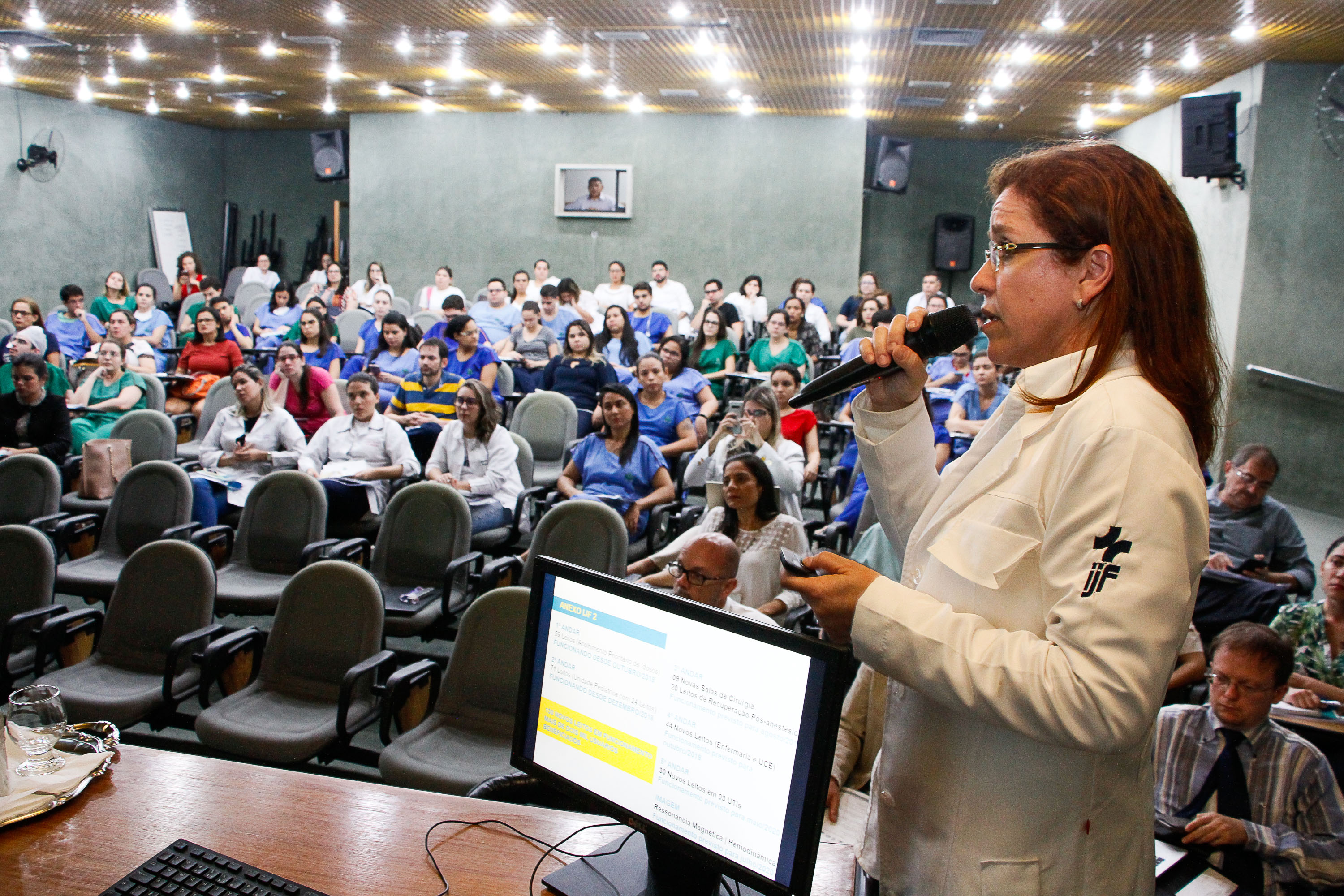 Mulher fala ao microfone para pessoas sentadas em auditório