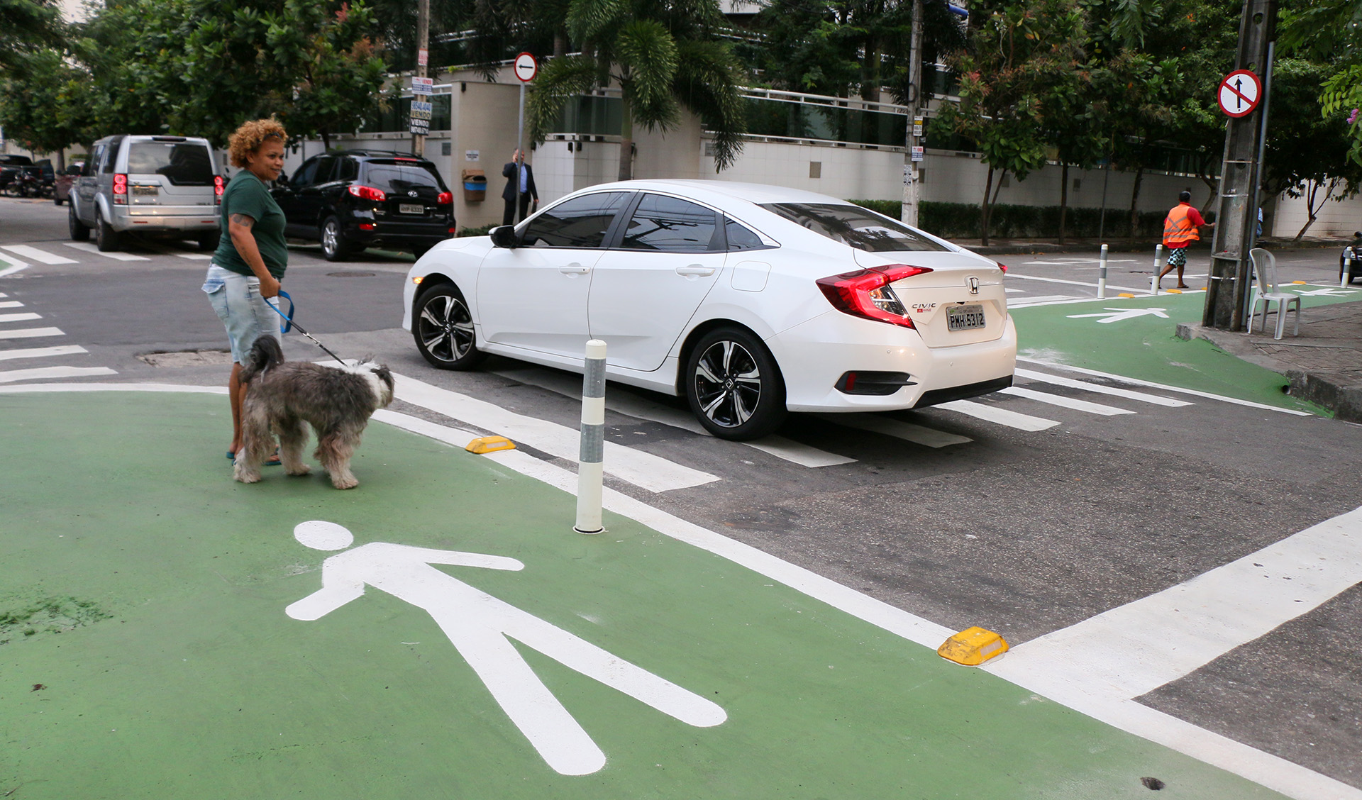 carro atravessa rua sinalizada e mulher com cachorro aguarda para atravessar