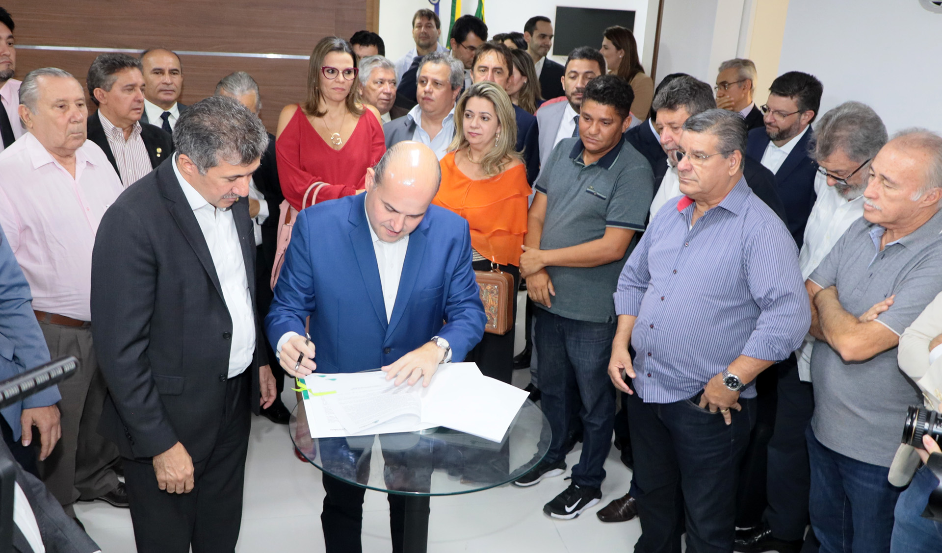 Prefeito assinando papel em cima de mesa e pessoas ao redor olhando