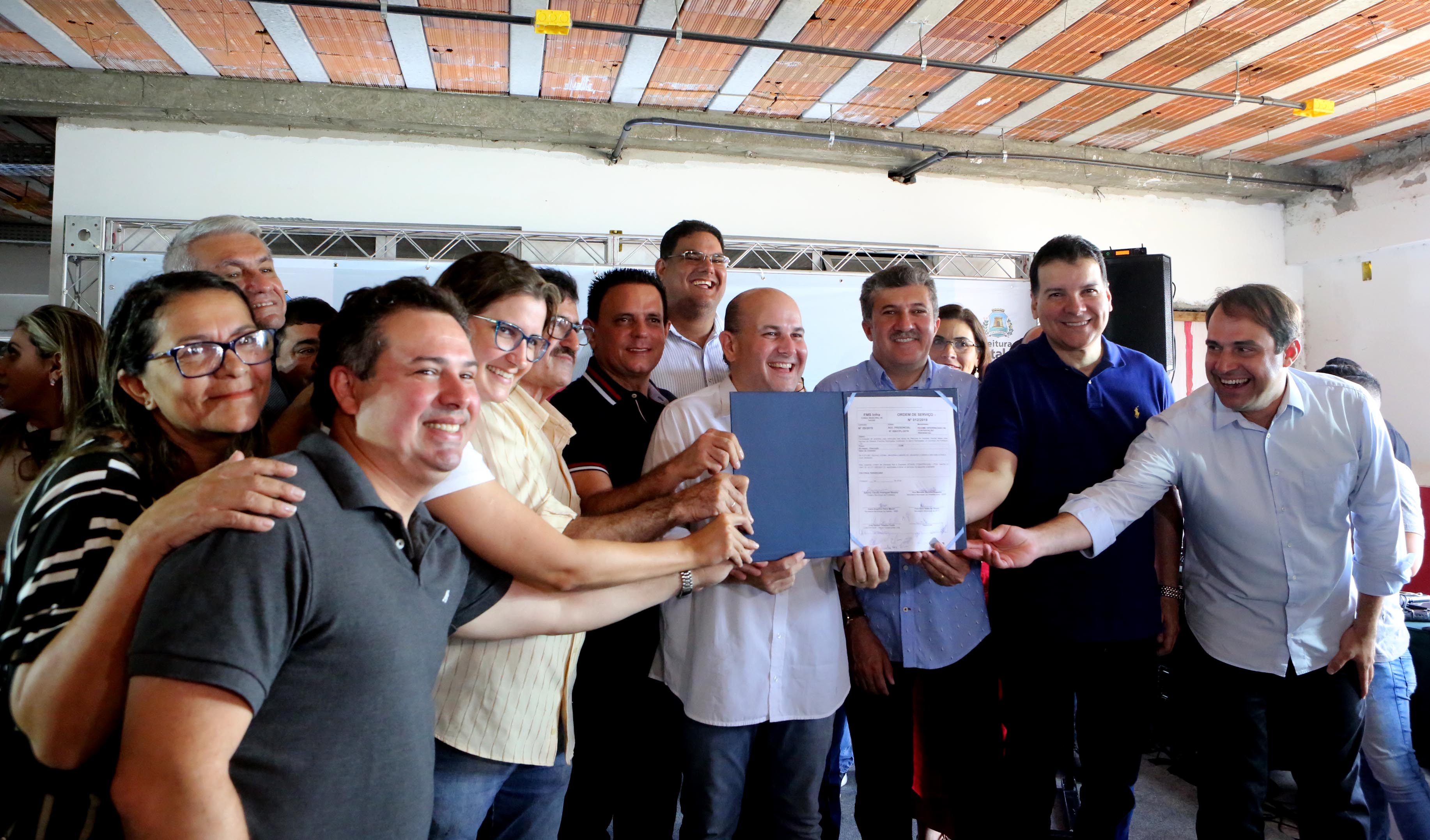 Prefeito ao centro sorrindo e segurando documento junto com pessoas ao redor