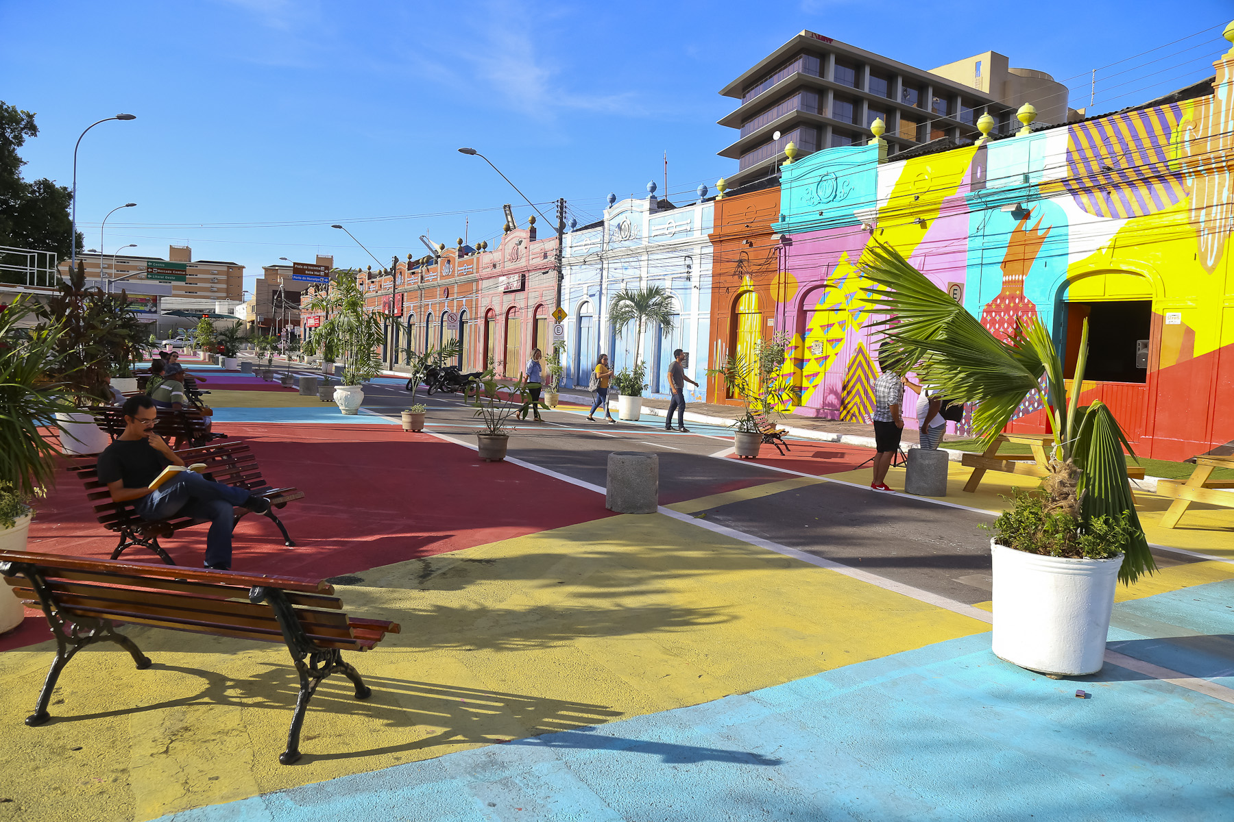 Ruas em frente ao Centro Dragão do mar com asfalto e imóveis pintados e coloridos, bancos de praça e pessoas sentadas