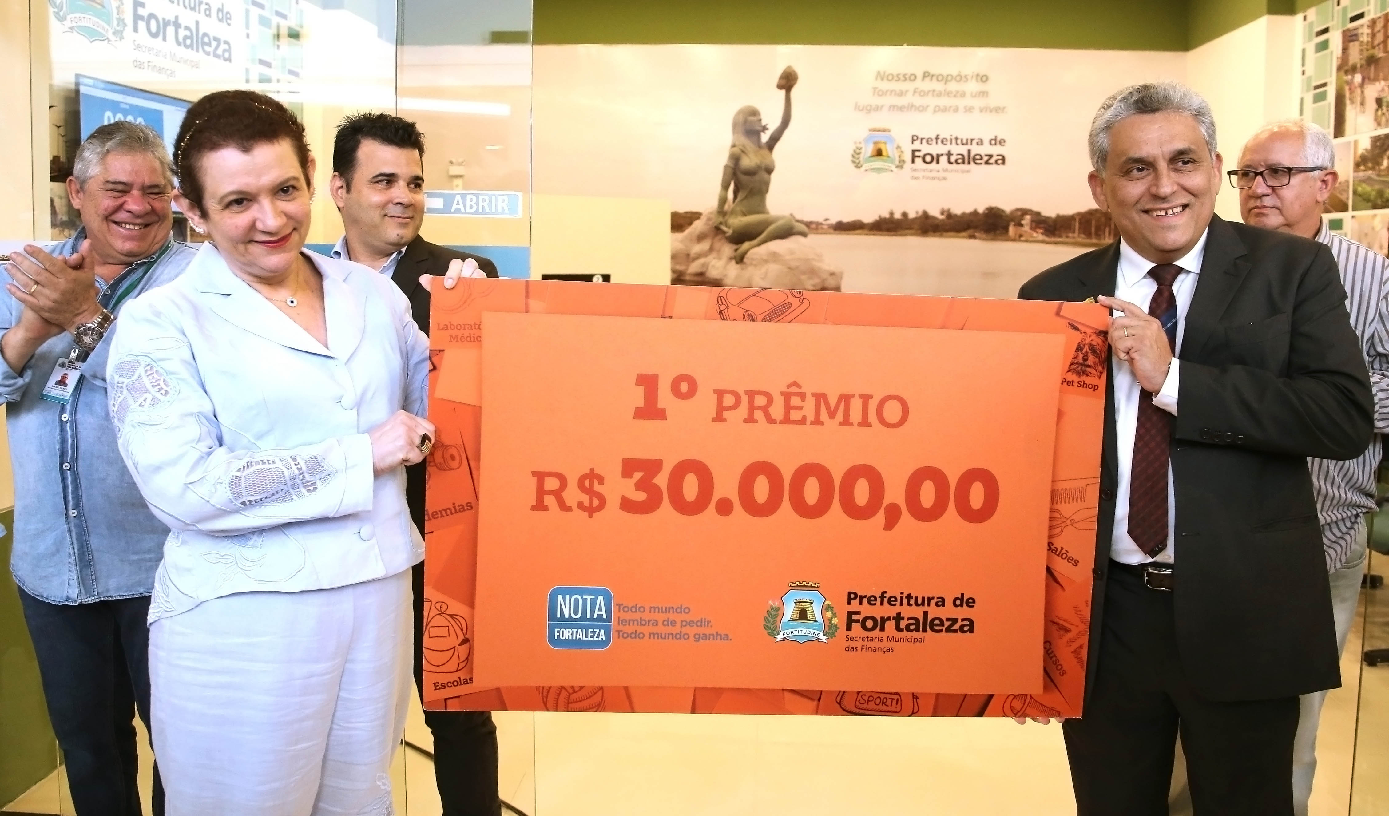 Mulher e homem segurando cartaz com valor de cheque de 30 mil reais e pessoas aplaudindo ao fundo