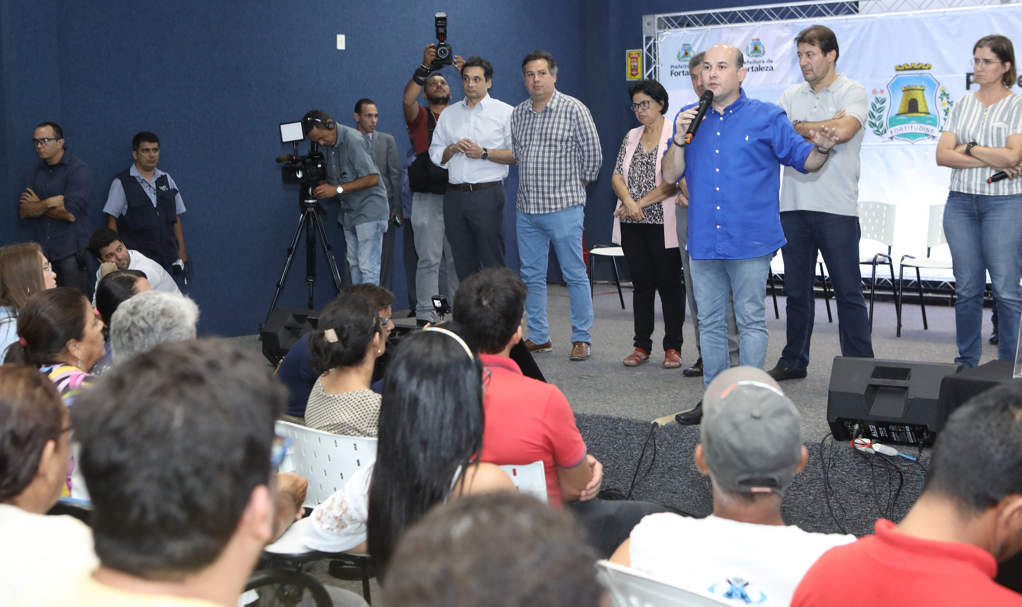prefeito, ao lado de secretários, falando ao microfone em cima de palco para pessoas na plateia sentada à frente