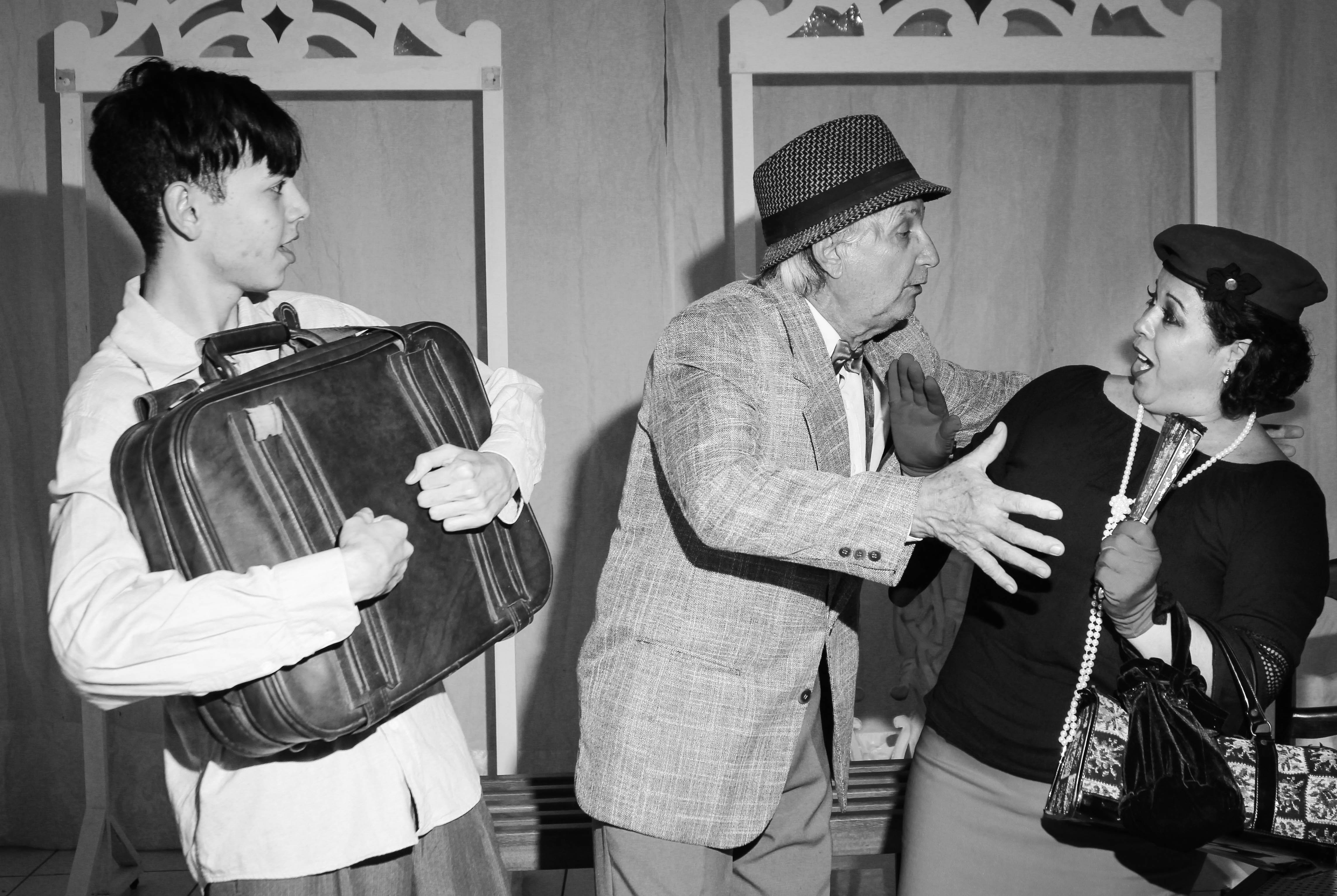Atores encenam espetáculo Os Coriscos: um jovem segura uma mala ao peito, enquanto um velho de chapéu e roupas antigas se exalta com um jovem senhora que o repele