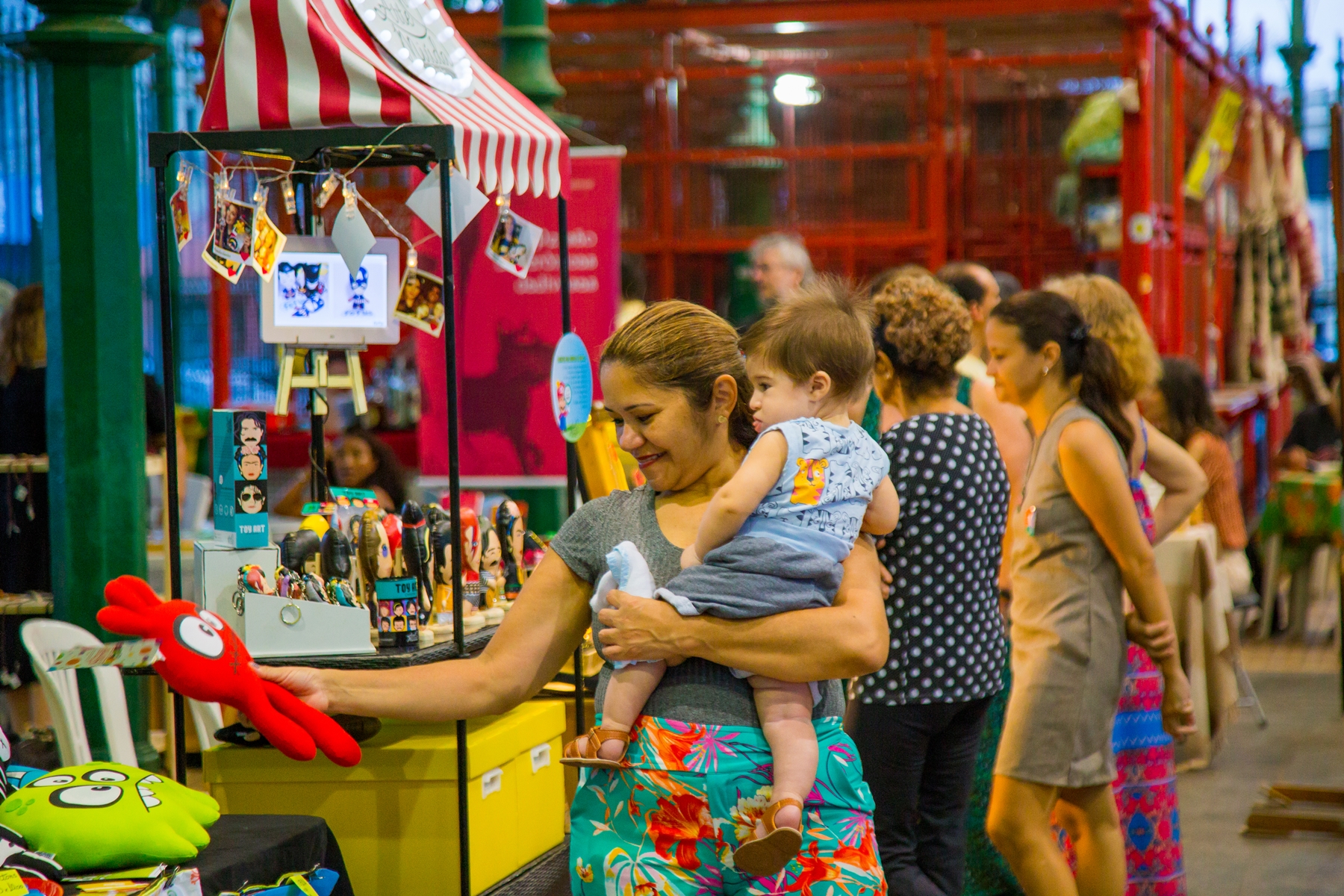 Mulher com criança no colo mostra brinquedos à venda no Mercado Coletivo