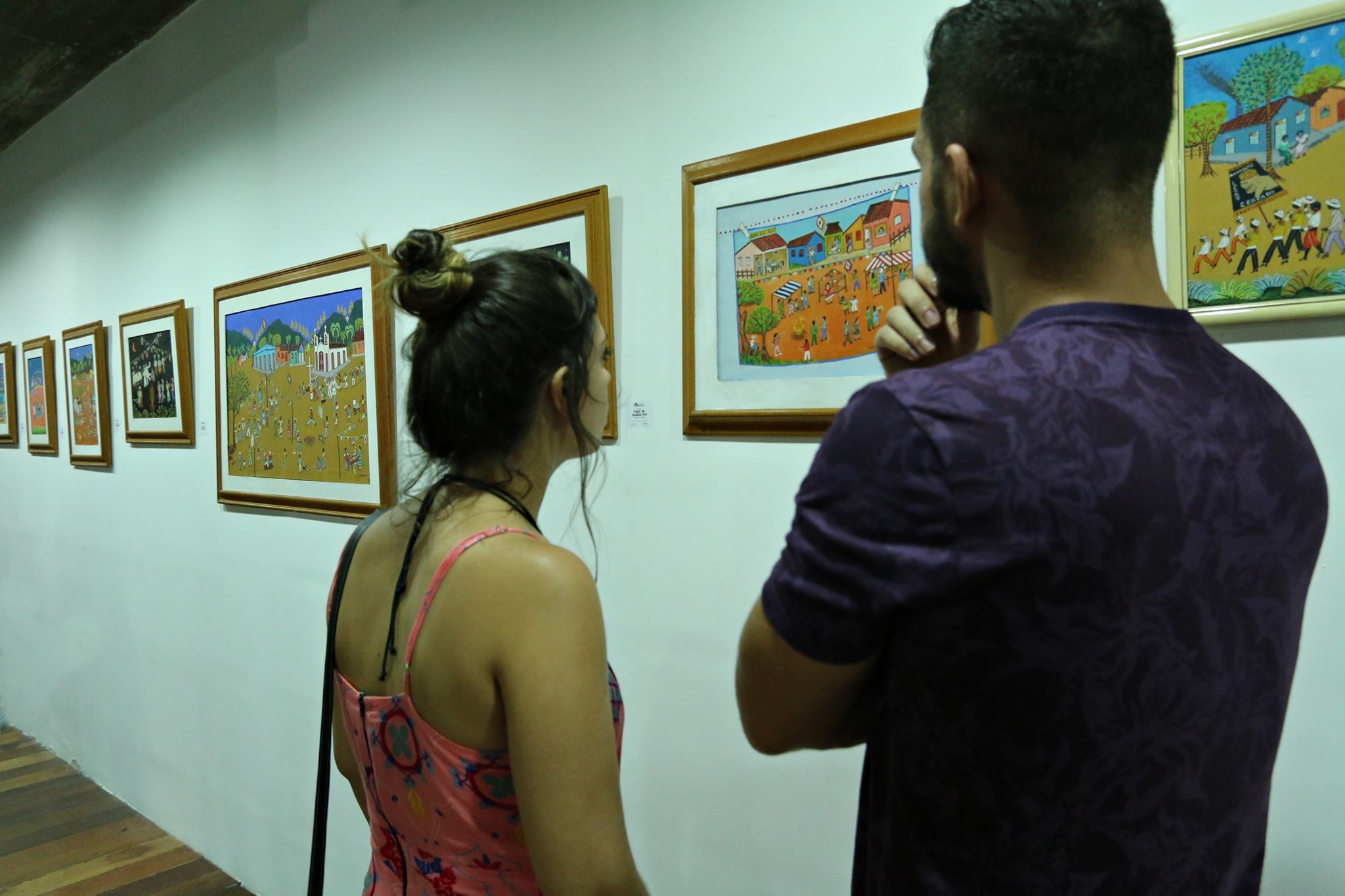 Pessoas olham quadros da exposição “Folias da Saudosa Rua” de Carmelita Fontenelle