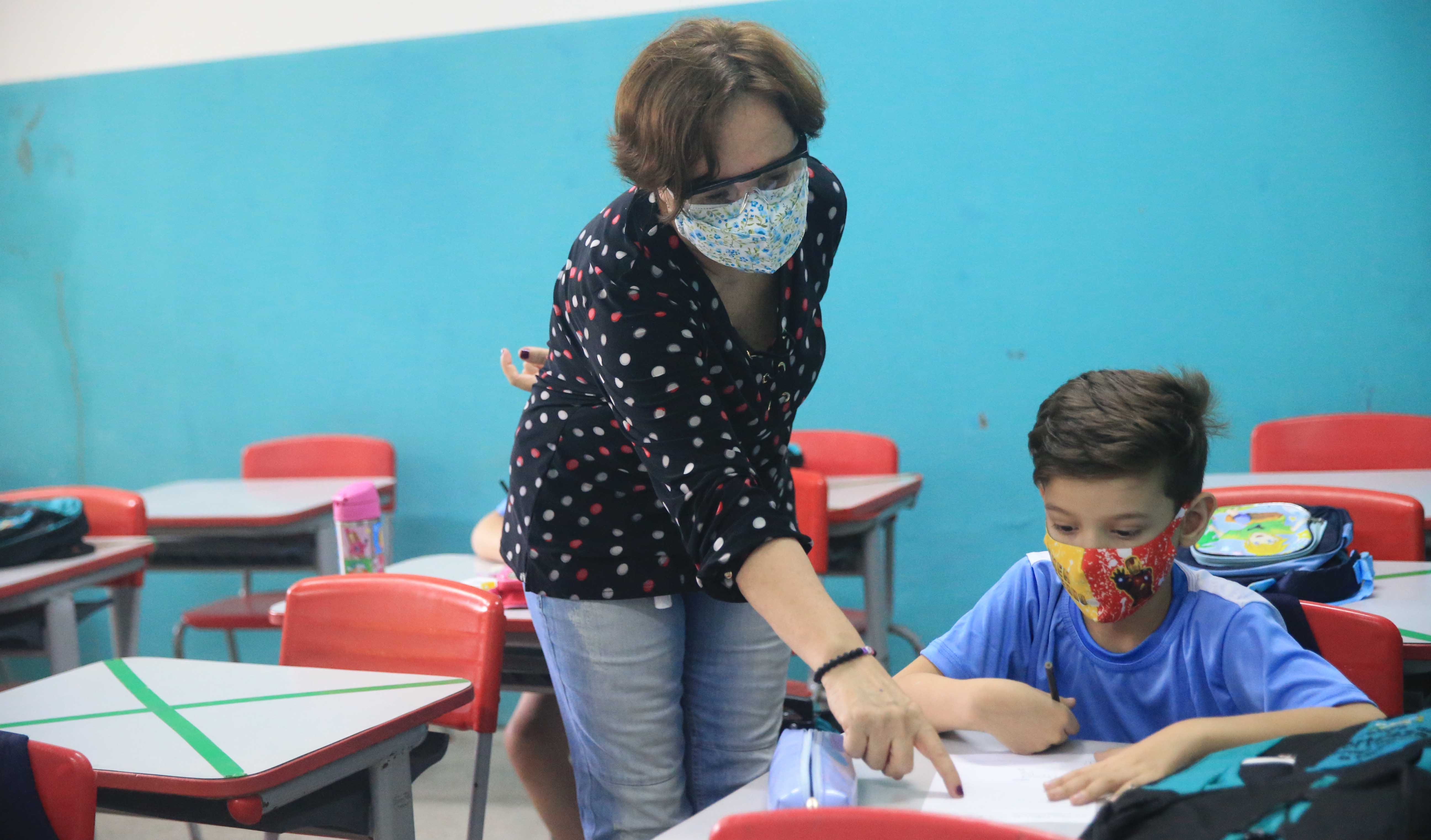 professor e aluno em sala de aula, ambos usam máscara e aluno lê um livro observado pela professora