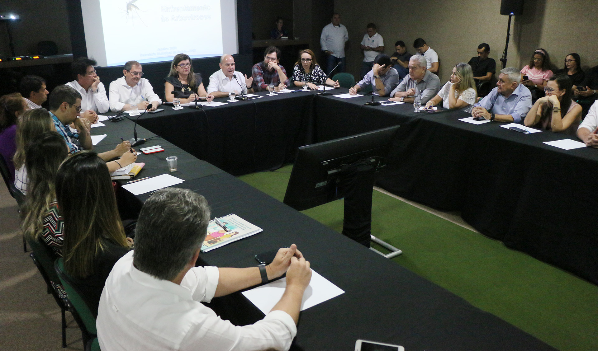 prefeito e secretária sentados em mesa com pessoas sentadas ao redor