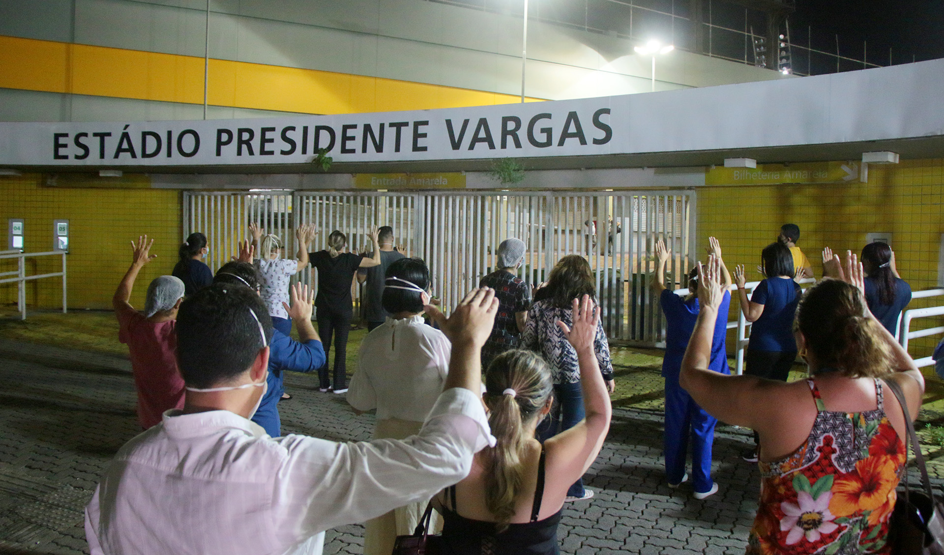 grupo de pessoas de costas com mãos para cima e cabeça baixa em frente a portão com placa escrito Estádio Presidente Vargas