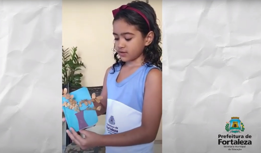 garota vestindo farda da rede municipal de ensino mostrando trabalho feito à mão e olhando para ele