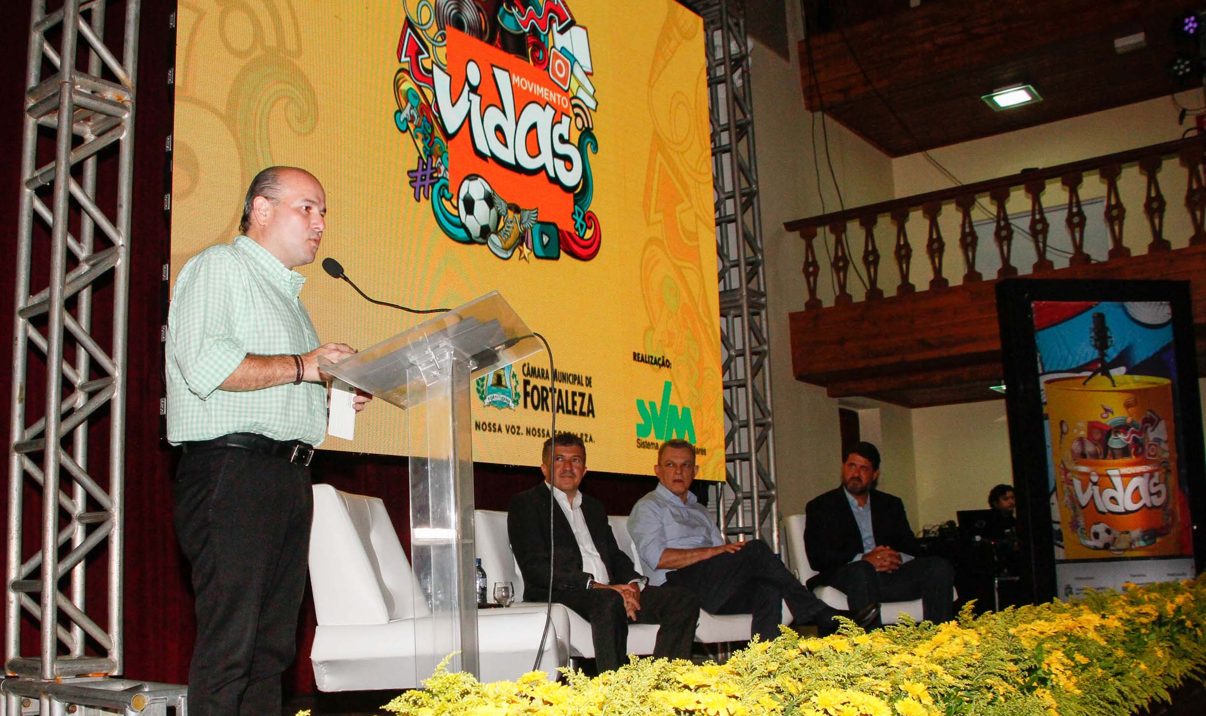 prefeito falando ao microfone em cima de palco e três senhores sentados ao lado