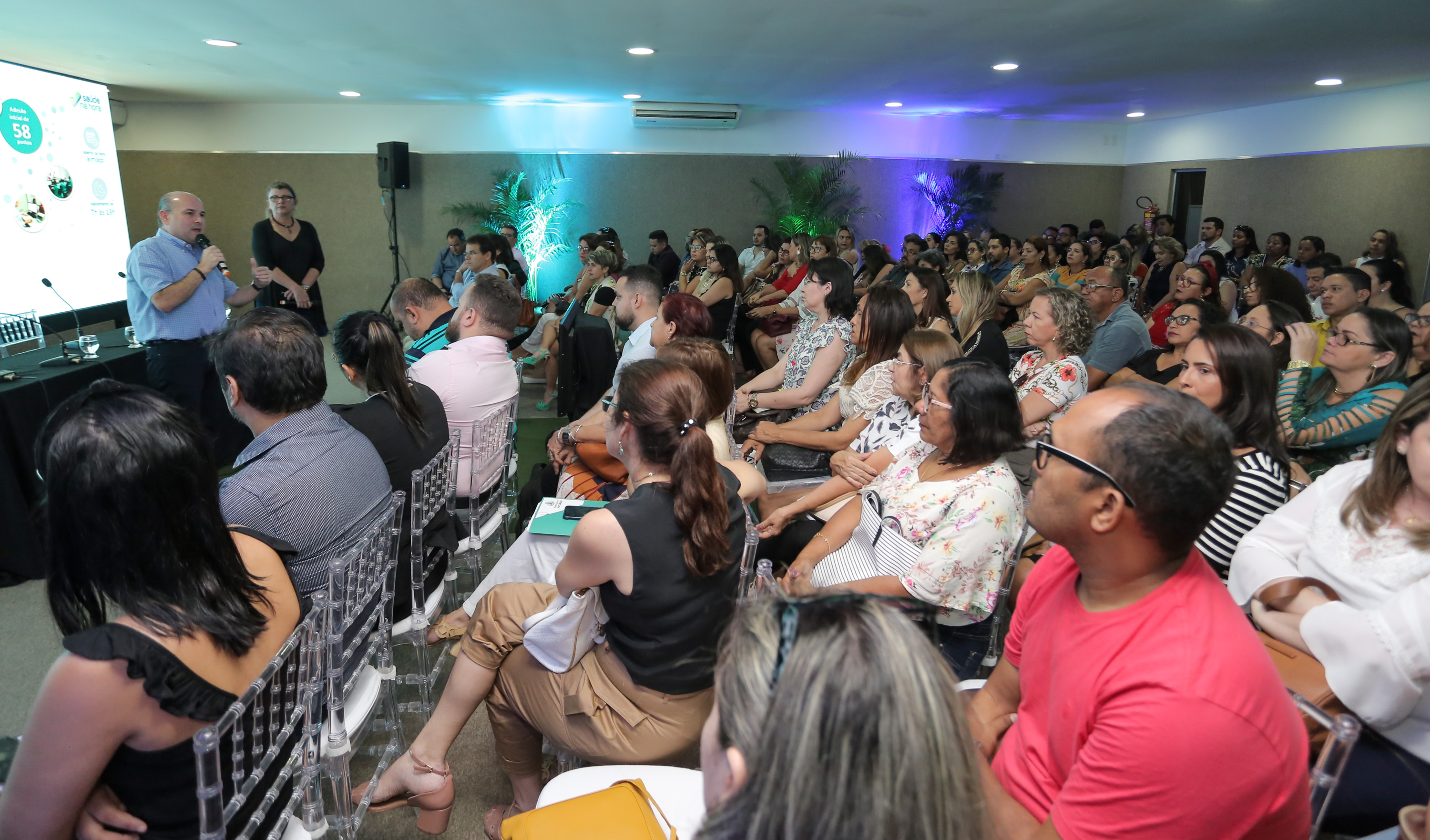 Prefeito, ao lado da secretária, em pé falando ao microfone para pessoas sentadas à frente