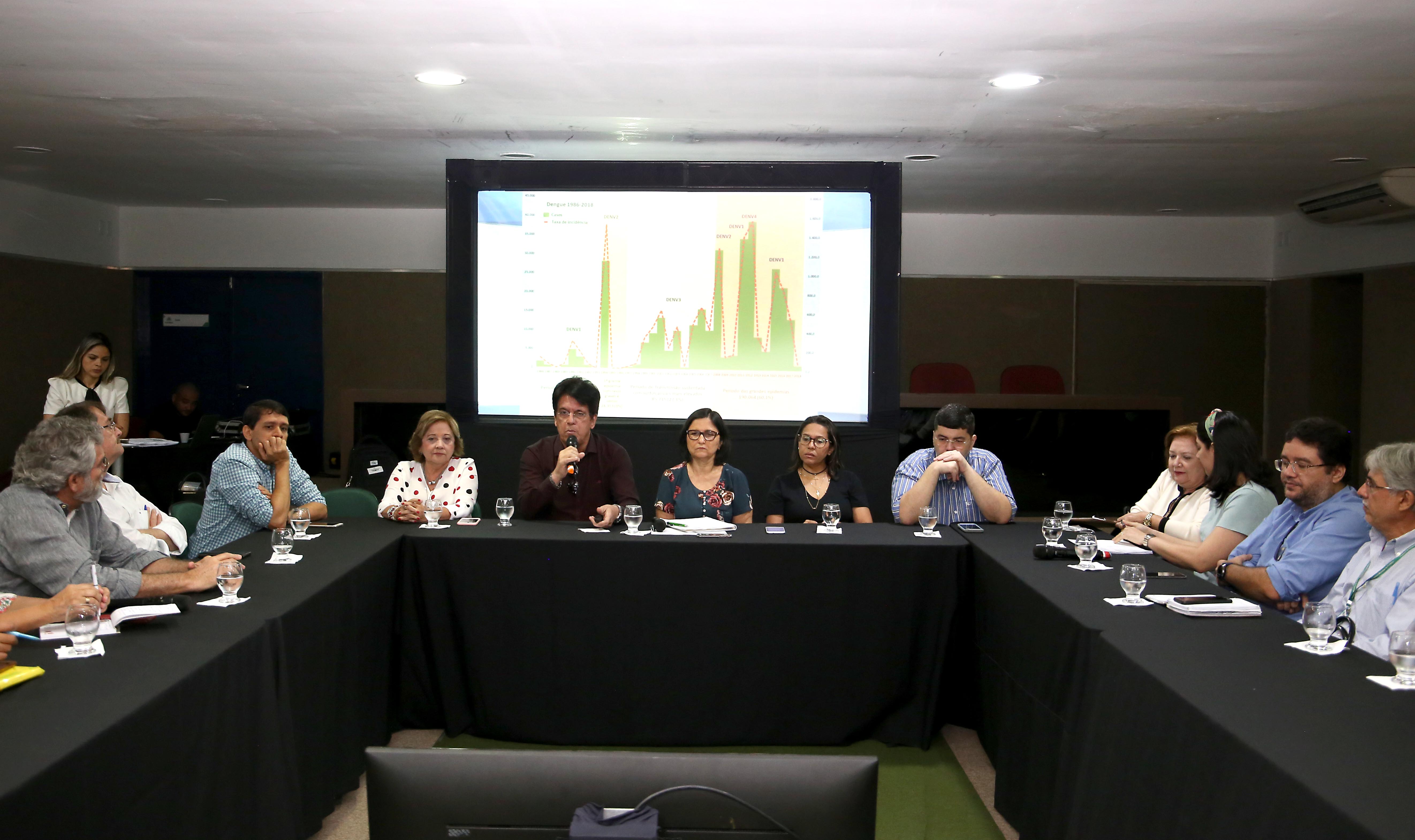 pessoas reunidas em mesa e homem falando em microfone ao centro