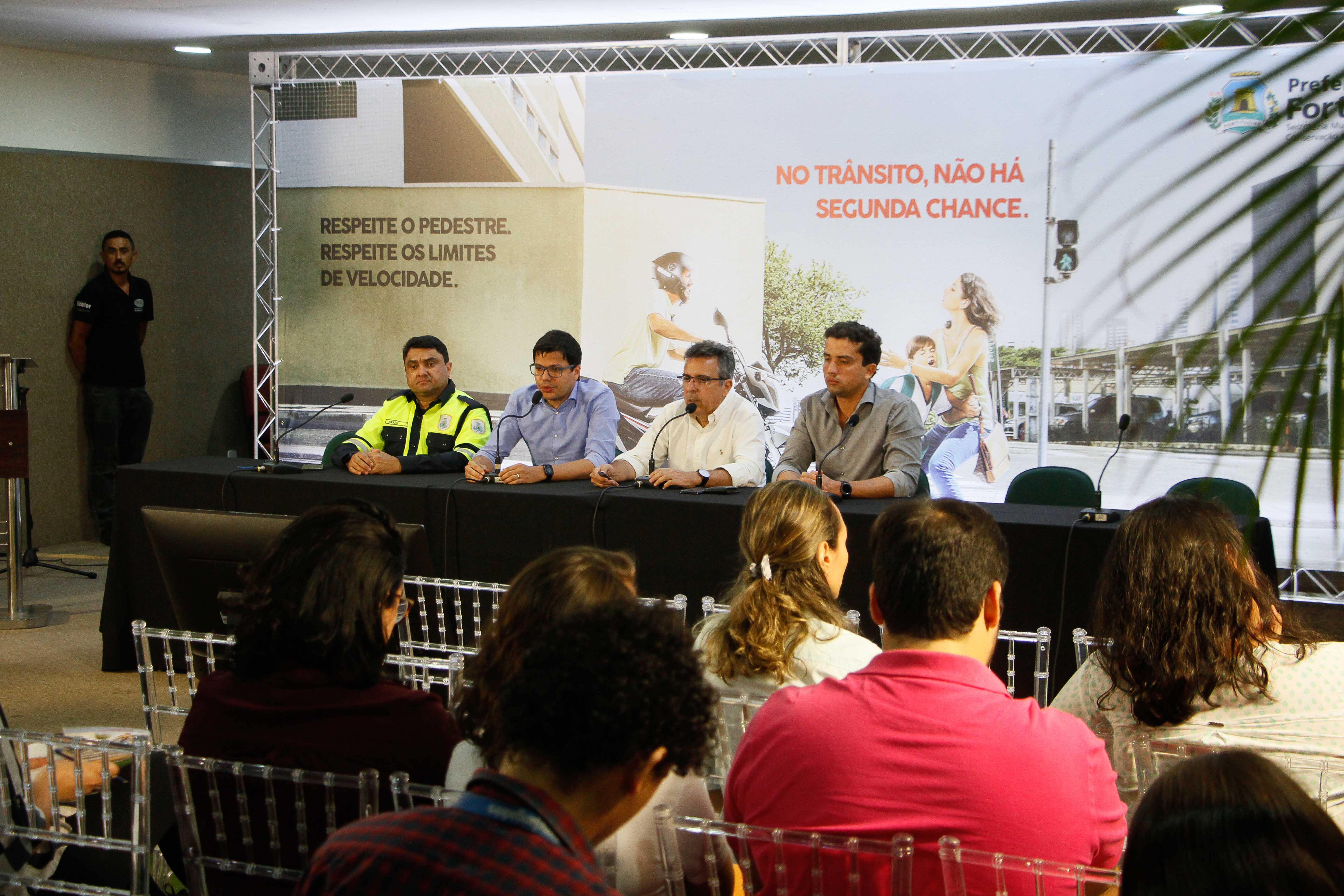 homens sentados em mesa falando para plateia sentada em frente