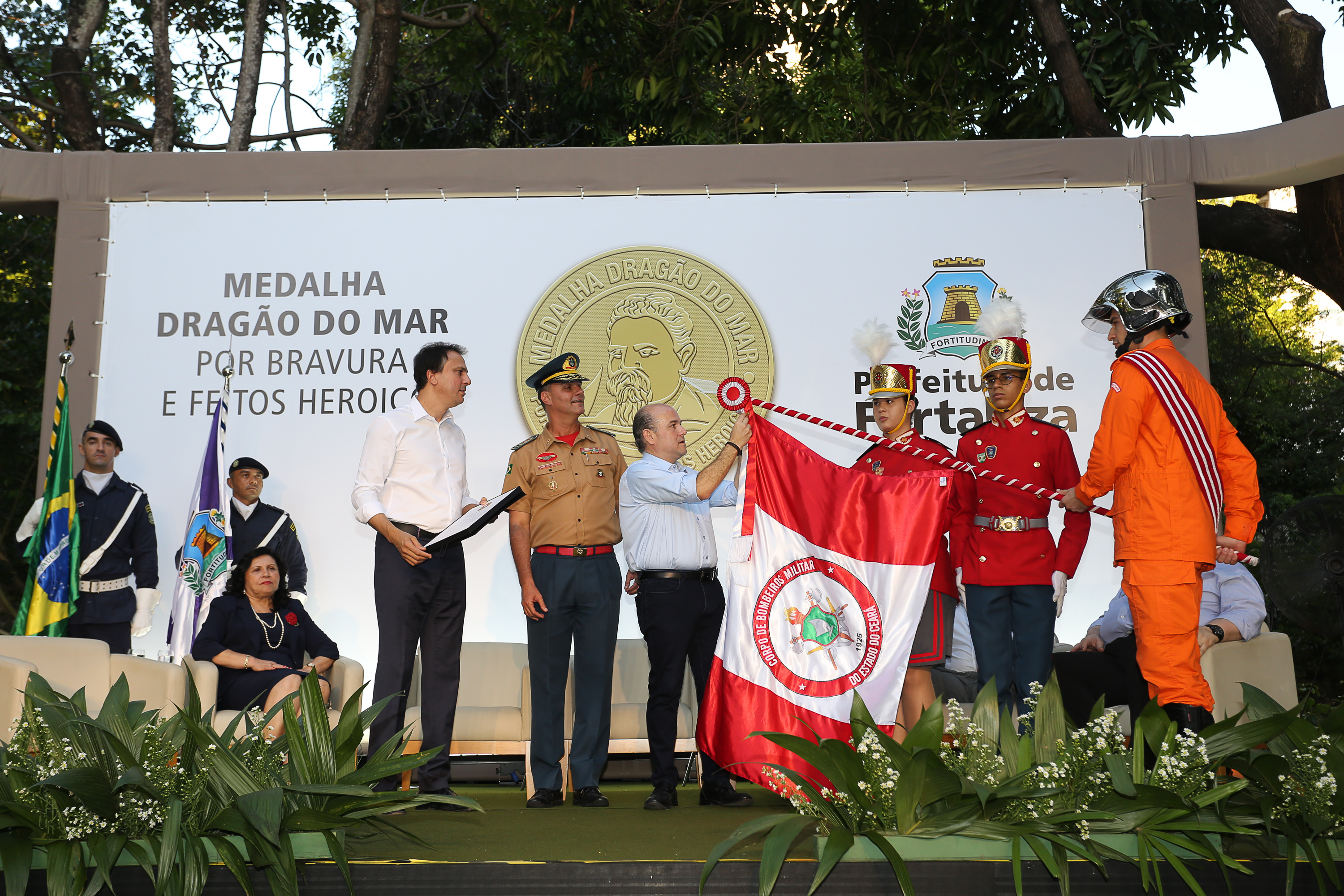 prefeito em cima de palco colocando medalha em bandeira com pessoas ao lado assistindo