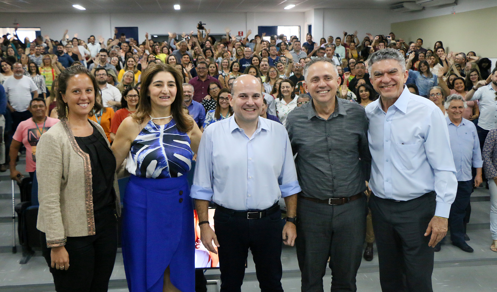 prefeito em pé com secretários em cima de palco com público ao fundo posando pra foto