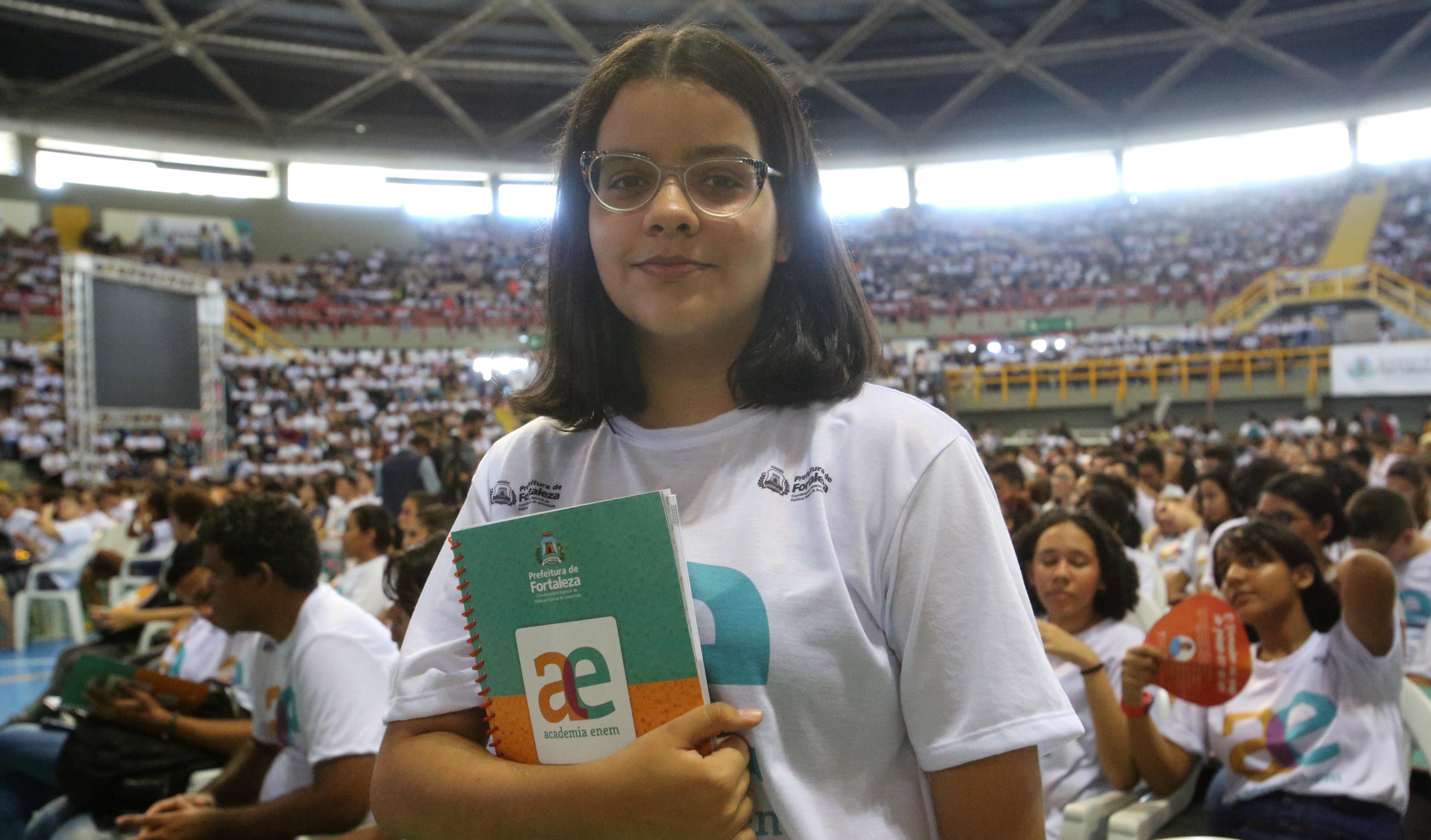 garota adolescente posando para a foto usando camiseta da Academia Enem e várias pessoas ao fundo no ginásio Paulo Sarasate