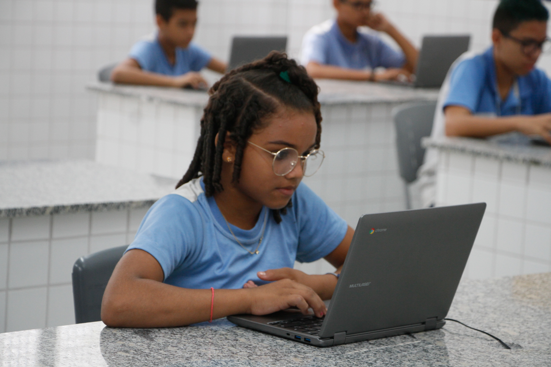 garota de óculos e farda da rede municipal sentada olhando para notebook e outras três crianças fazendo o mesmo em mesas ao fundo