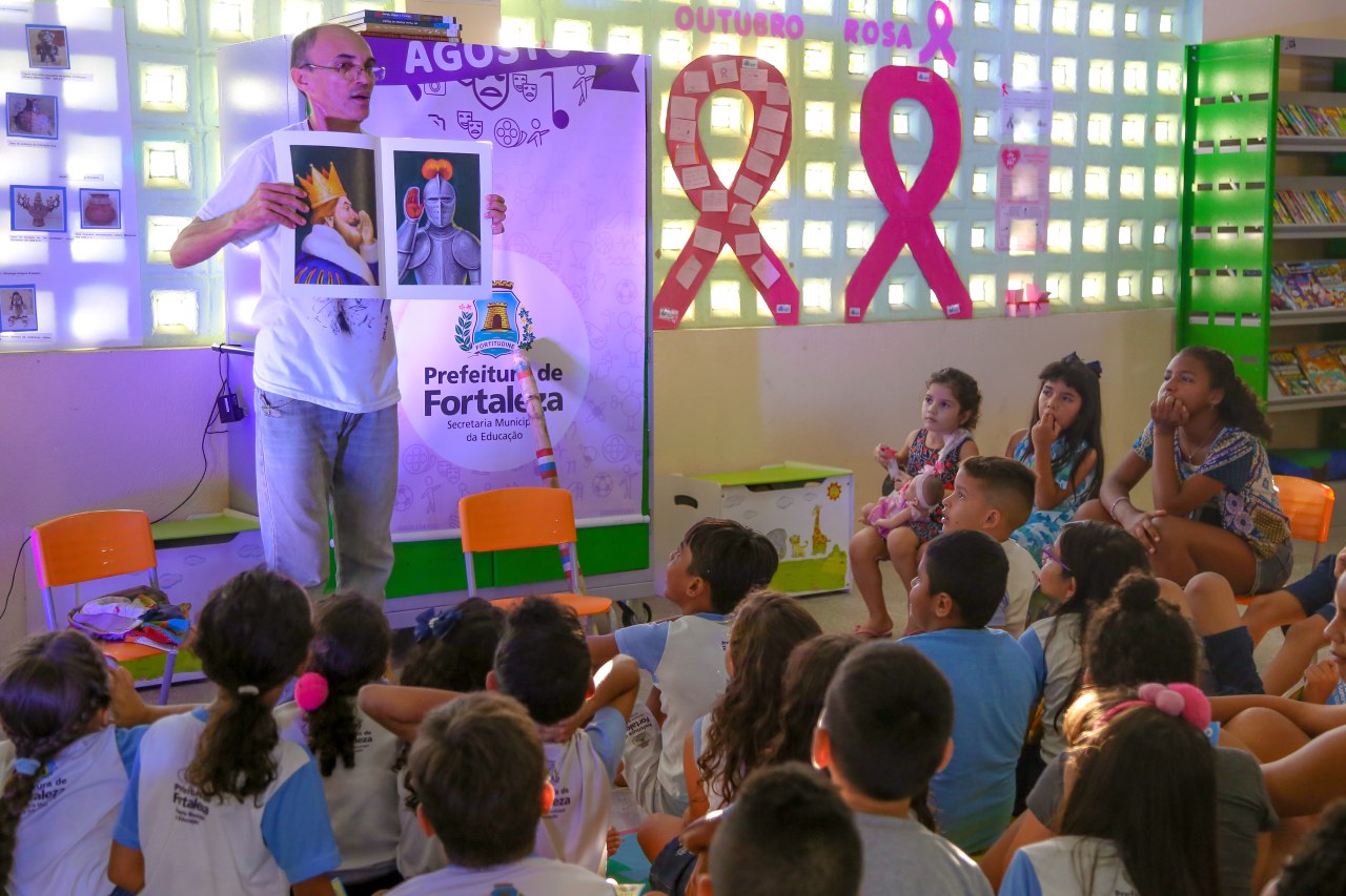 Contador de histórias Chicão Oliveira mostra um livro infantil em frente a crianças sentadas no chão