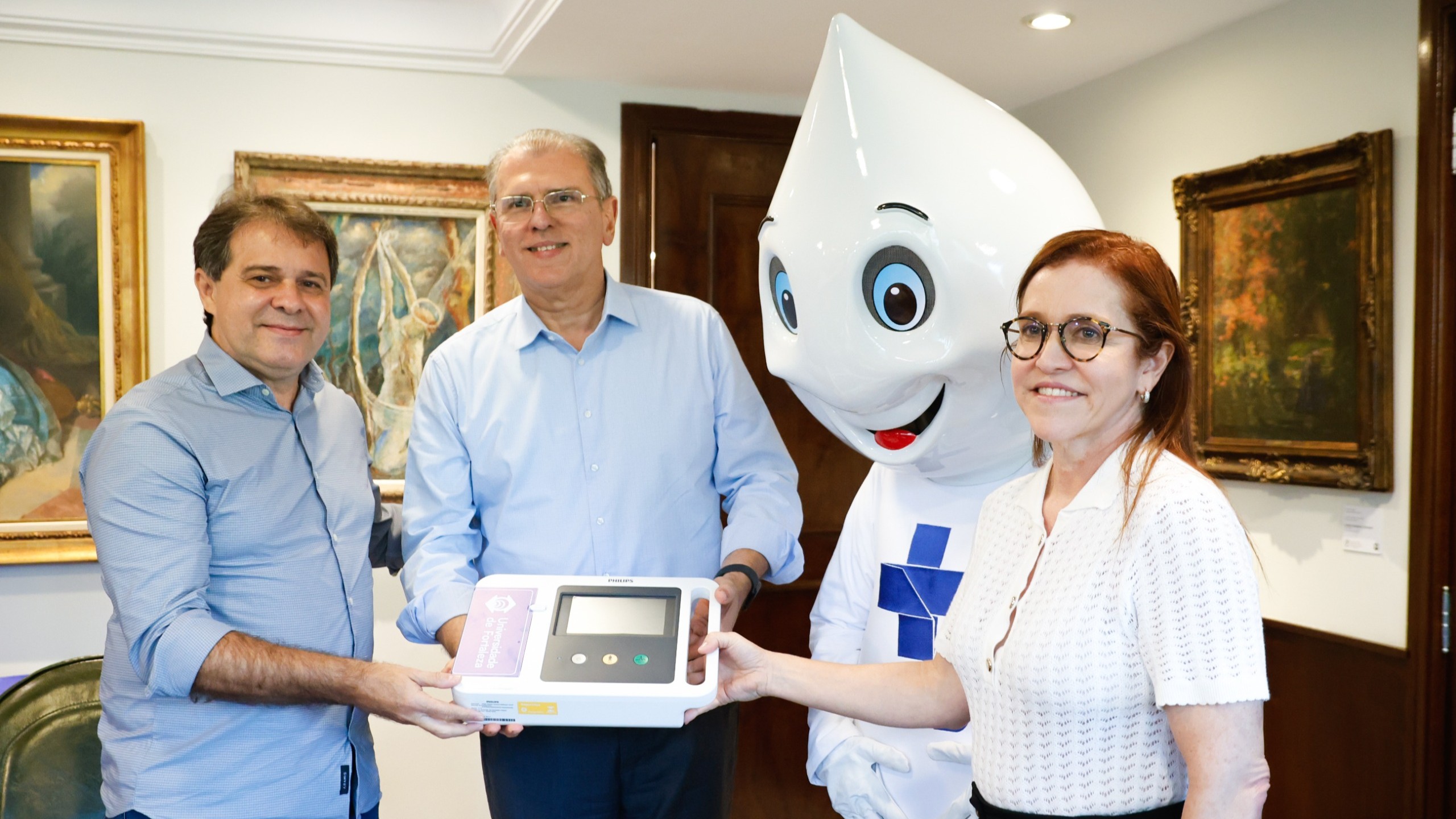 Prefeito Evandro Leitão, o reitor da Universidade de Fortaleza (Unifor), professor Randal Pompeu, e a secretária municipal da Saúde, Riane Azevedo seguram um aparelho de eletrocardiograma. Ao lado dele também está o Zé Gotinha.