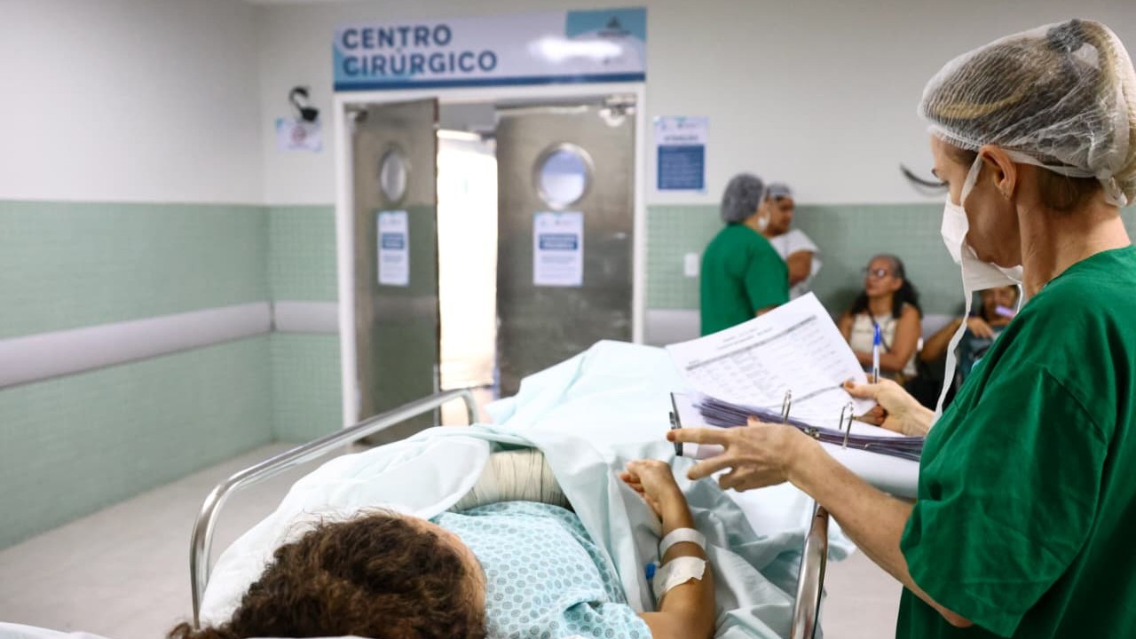 Paciente aguarda por uma cirurgia no IJF. Ela está deitada em uma maca, com uma enfermeira em pé ao seu lado, e em segundo plano, ao fundo, se vê a entrada do Centro Cirúrgico
