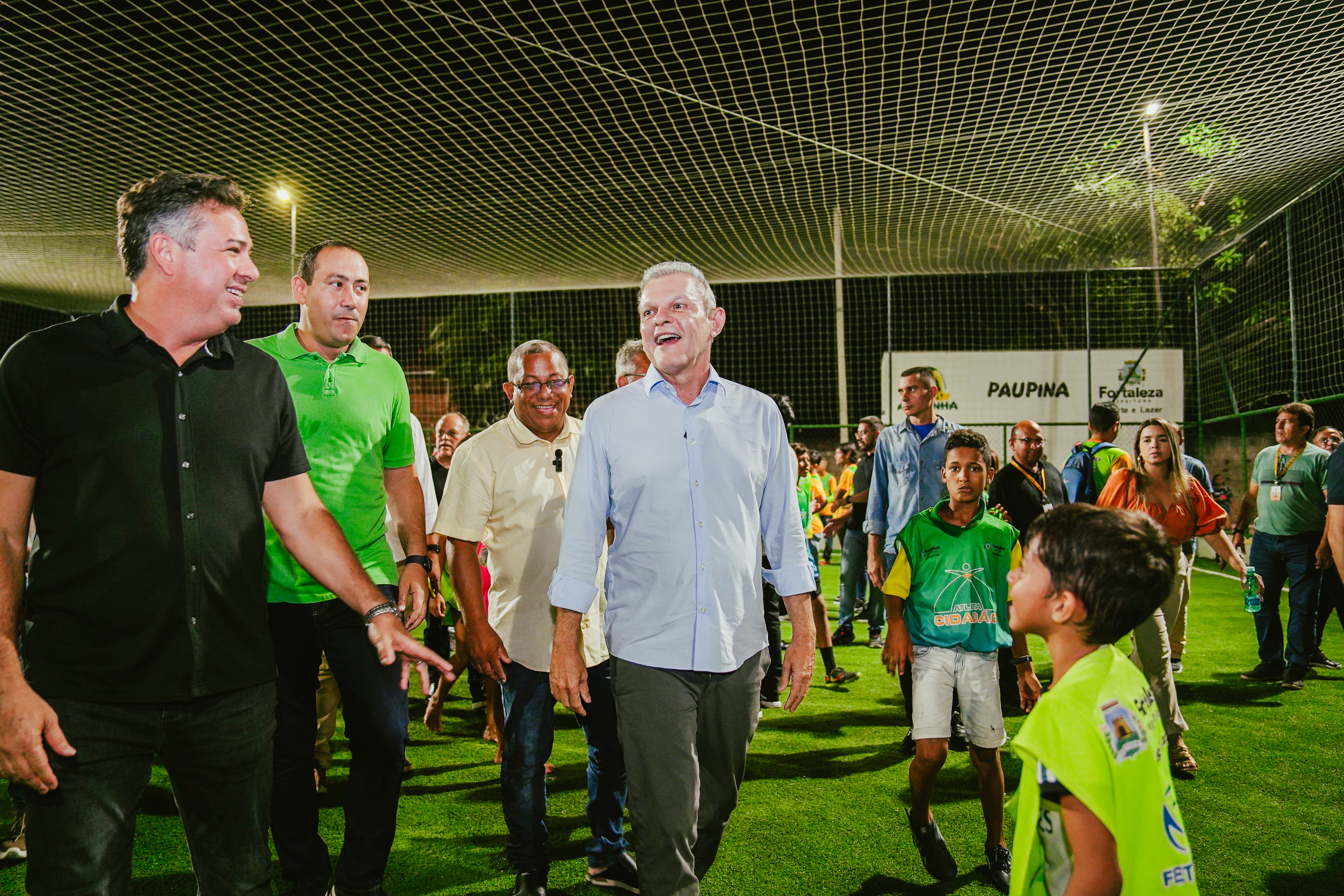prefeito sorrindo enquanto anda pelo gramado da areninha com adultos e crianças