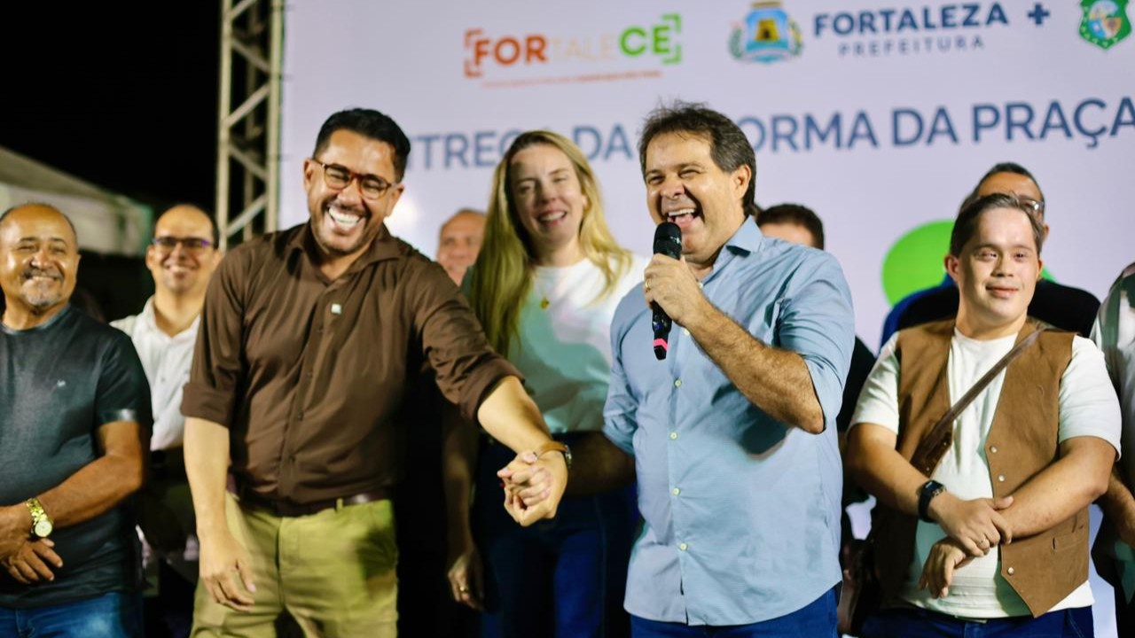A imagem mostra o palco do evento de entrega da praça. O prefeito Evandro Leitão e o vereador Márcio Martins estão em primeiro plano, rindo. 