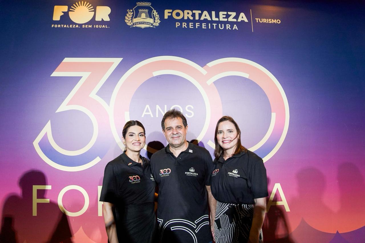 A foto mostra o prefeito Evandro Leitão, a primeira dama Cristiane Leitão e a secretária do Turismo Denise Carrá posando em frente ao backdrop dos 300 anos de Fortaleza