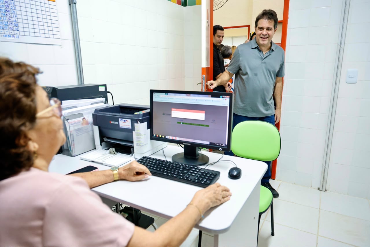 O prefeito Evandro Leitão entra em uma das salas do posto de saúde recém inaugurado. Em primeiro plano há uma servidora do posto usando um computador