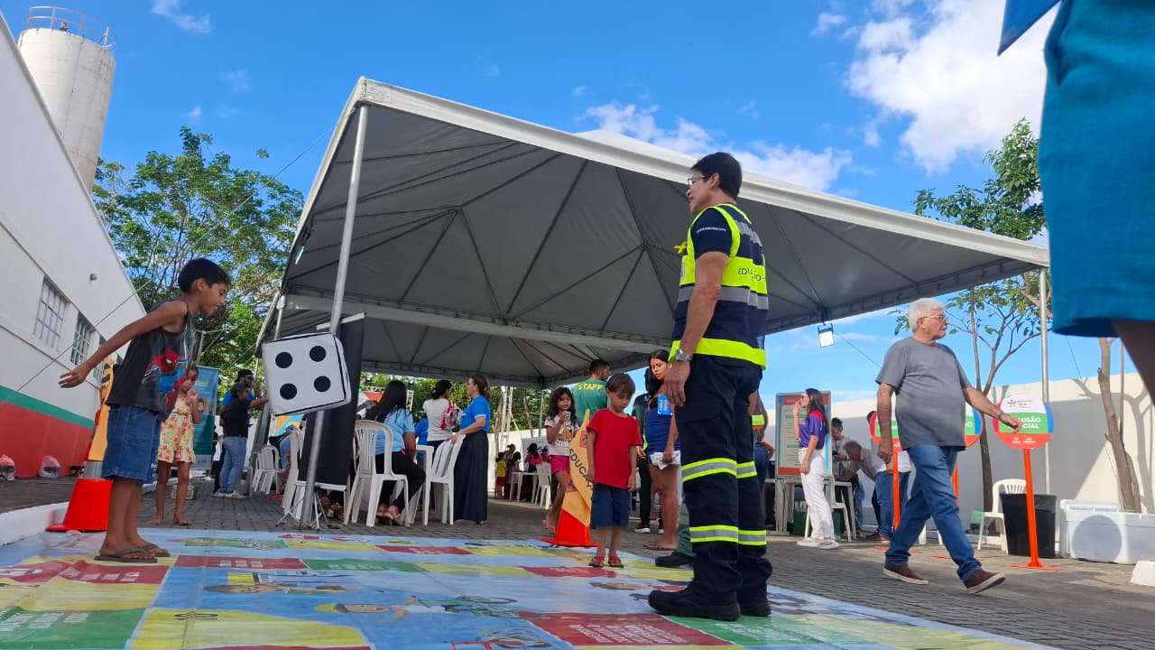 A foto mostra uma criança jogando um dado em cima de um jogo de tabuleiro gigante. Ao fundo aparecem as tendas das ações do projeto Infância Viva na Praça