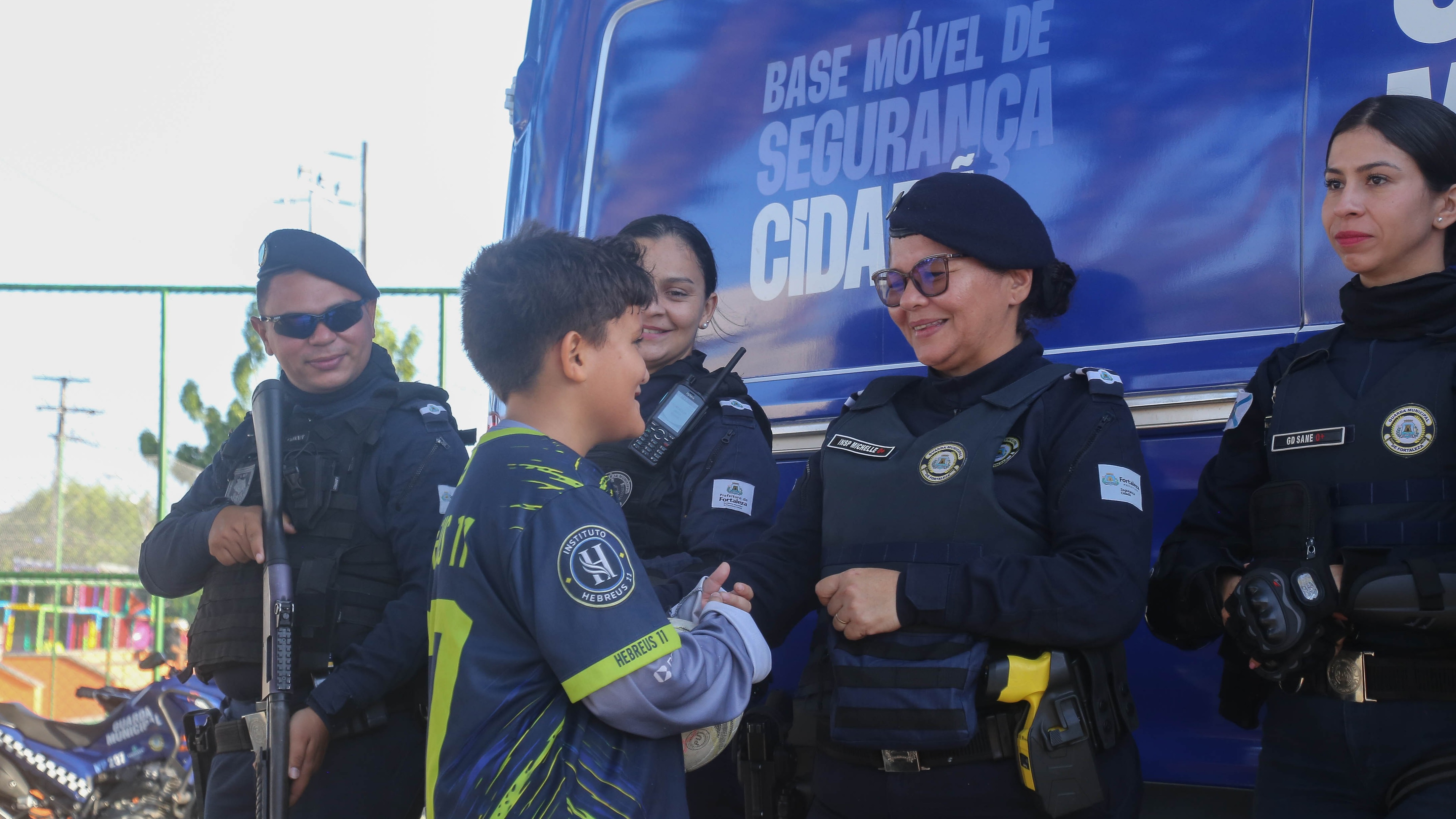 Guardas municipais estão em frente a uma base móvel dos Pelotões dos Bairros. Uma das guardas está cumprimentando uma criança