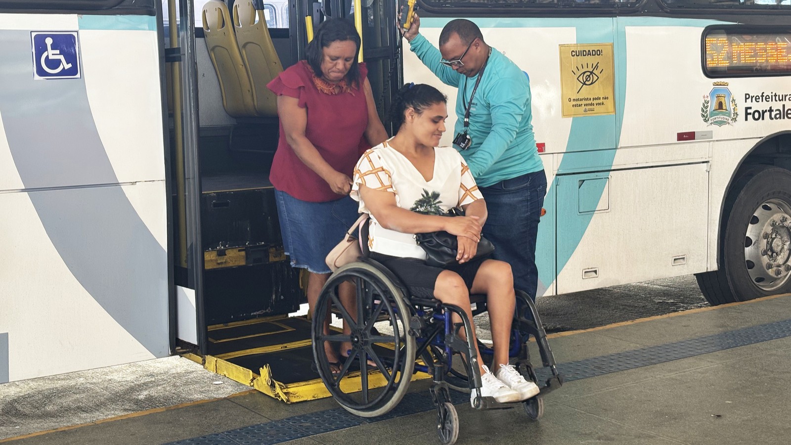 a foto mostra uma mulher numa cadeira de rodas acessando um ônibus pela plataforma, auxiliada pelo motorista e por uma acompanhante