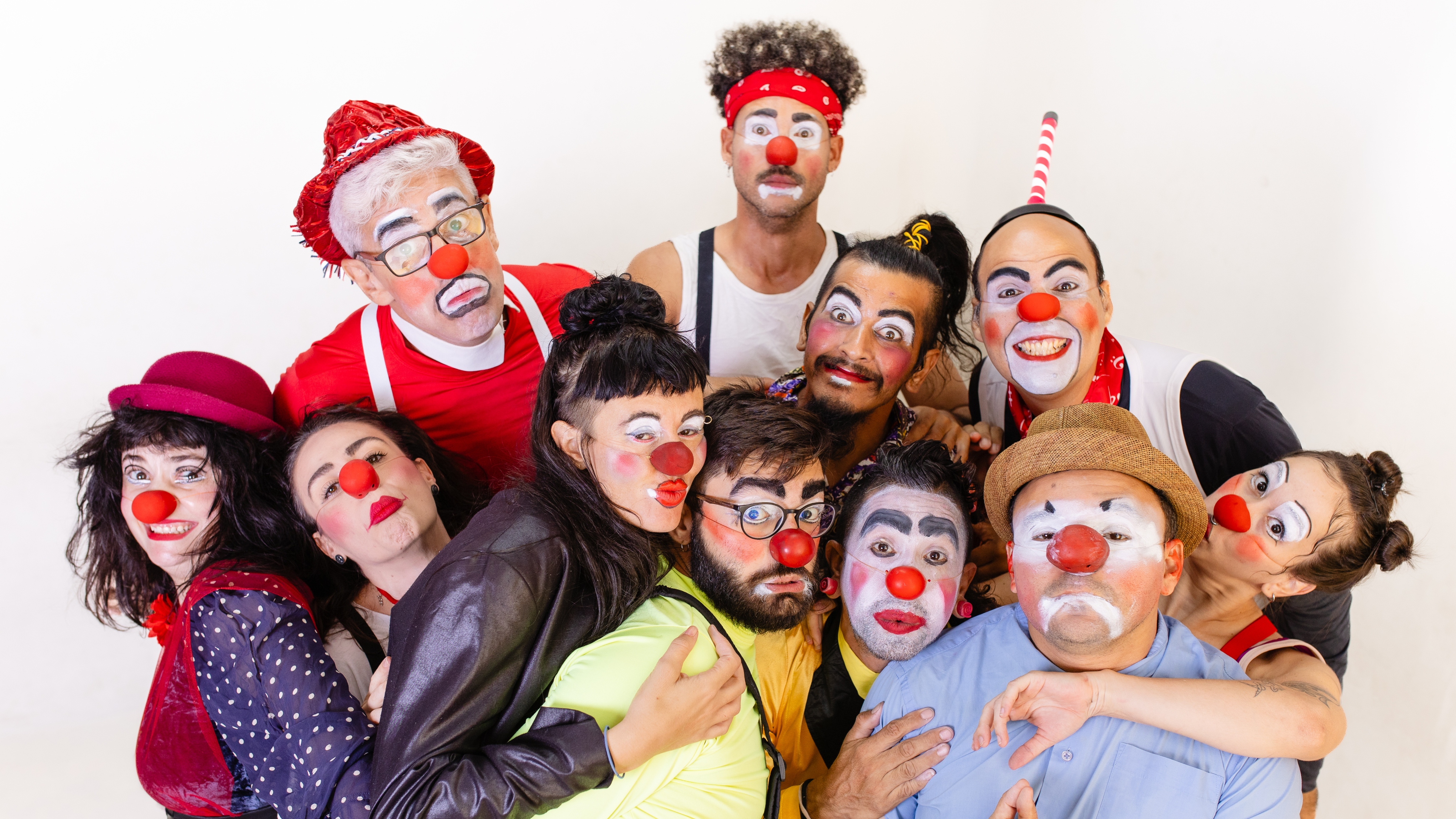 Fotografia em grupo de dez pessoas caracterizadas como palhaços, com narizes vermelhos e maquiagens faciais expressivas. Elas posam juntas, sobrepostas, olhando para a câmera contra um fundo branco.